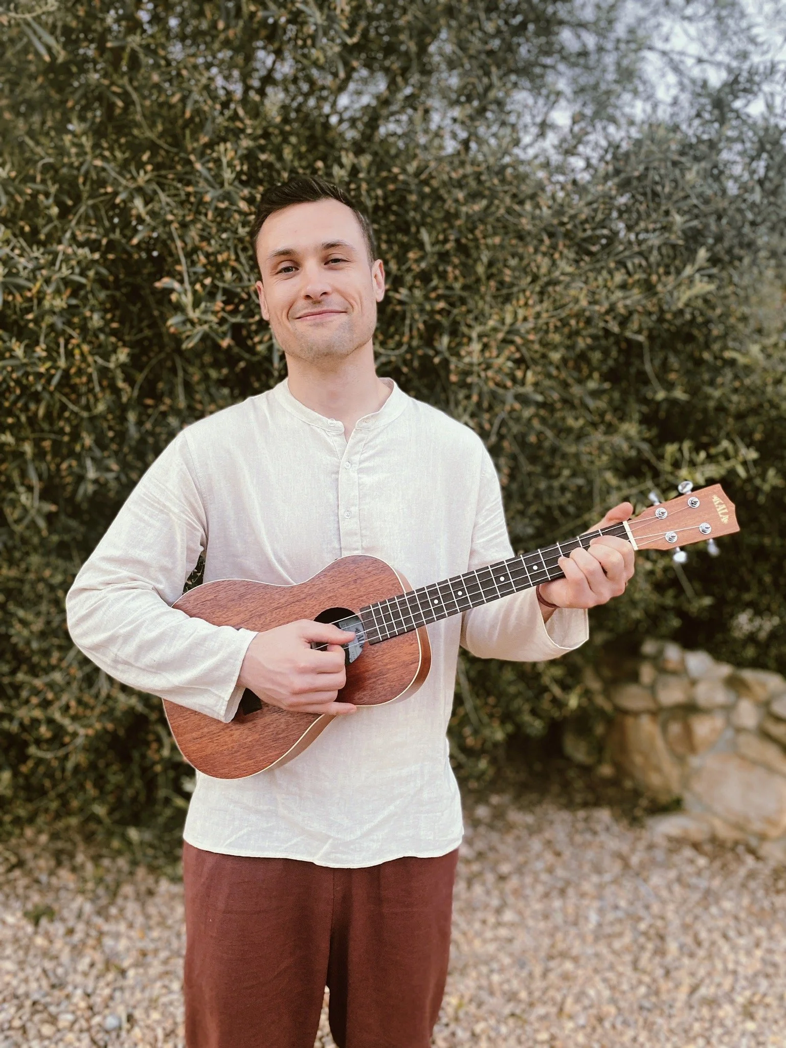 A young man is standing outdoors in front of green bushes, holding a small wooden ukulele, and smiling at the camera.