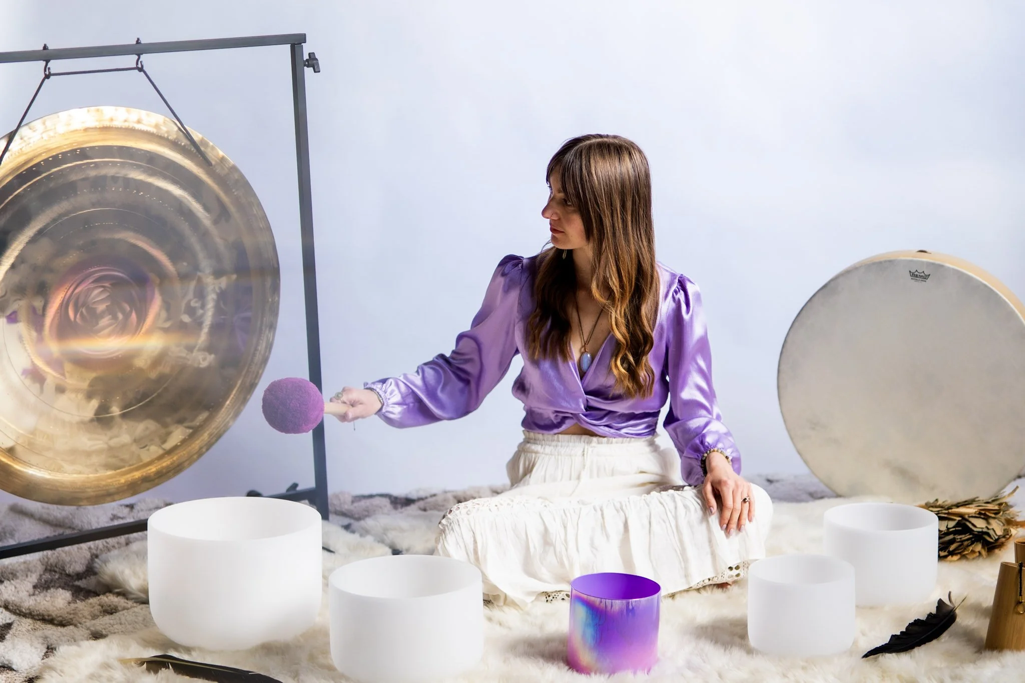 A woman sitting on a soft, plush surface, wearing a purple satin top and white skirt, playing a large singing bowl with a mallet, surrounded by other colorful bowls, with a large gong on her left.