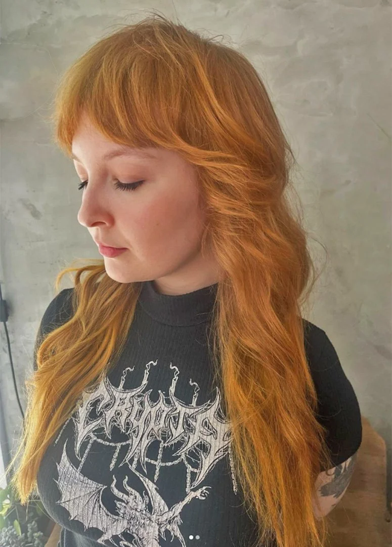 A woman with long, wavy red hair and fair skin looking down, wearing a black t-shirt with white graphic design.