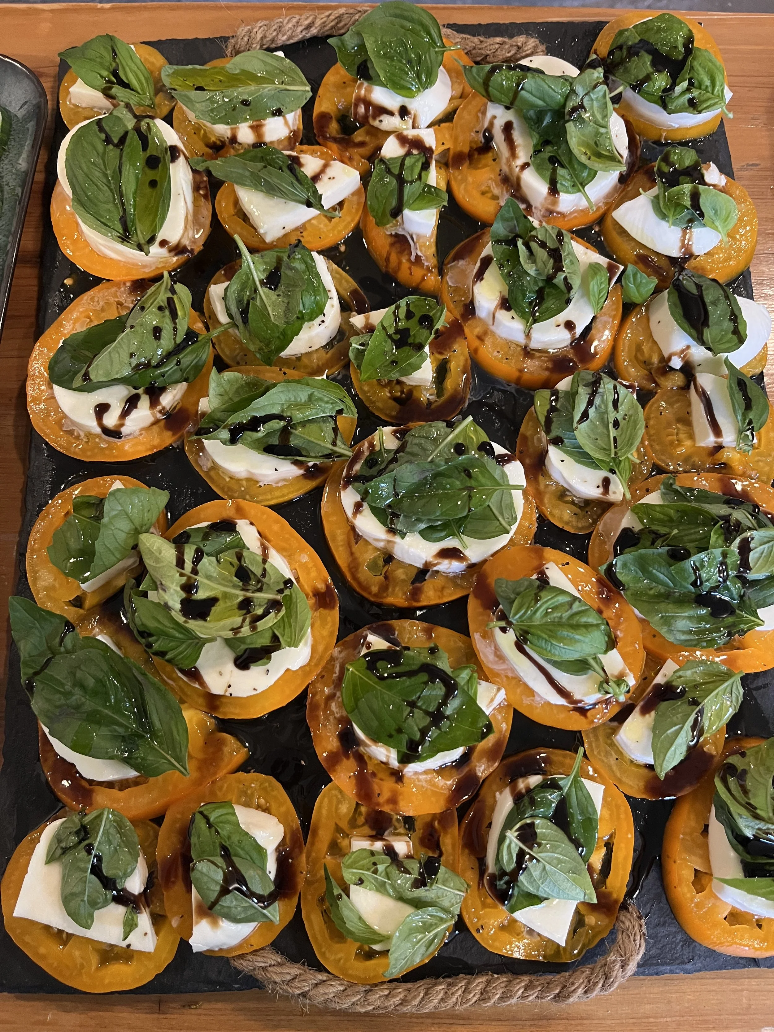 A black serving tray filled with sliced yellow and white heirloom tomatoes topped with fresh basil leaves, drizzled with balsamic glaze, and garnished with slices of fresh mozzarella cheese.
