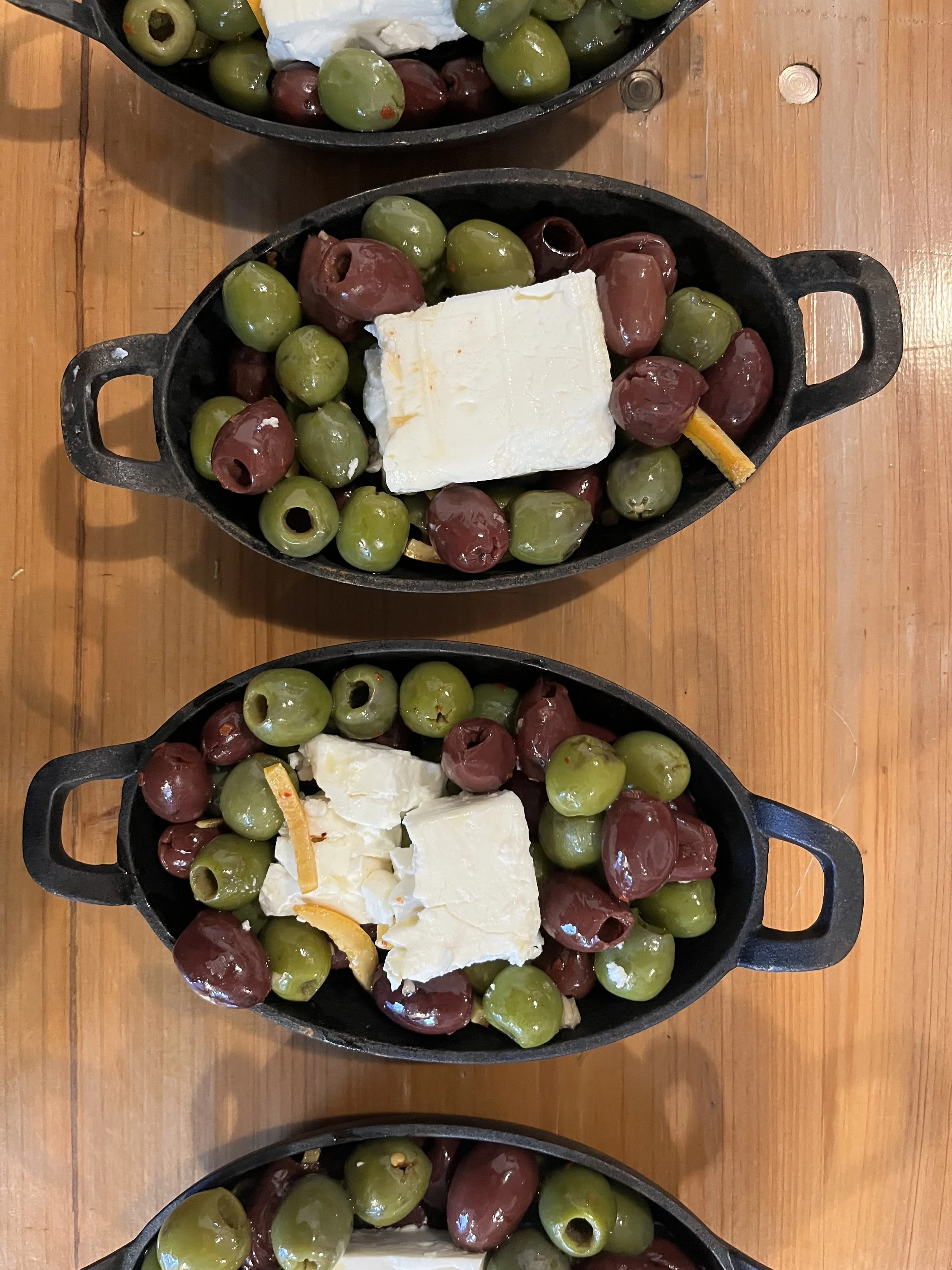 Cast iron skillet containing green and purple olives, several pieces of cheese, and a lemon peel atop a wooden table.