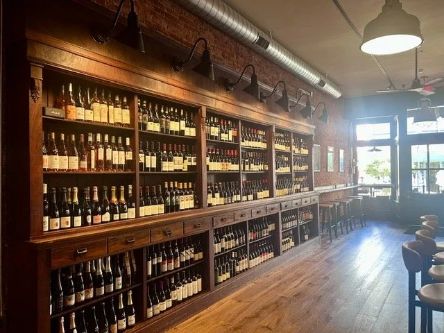Wine bottles on wooden shelves inside a cozy, well-lit wine shop or tasting room with a brick wall and large windows.