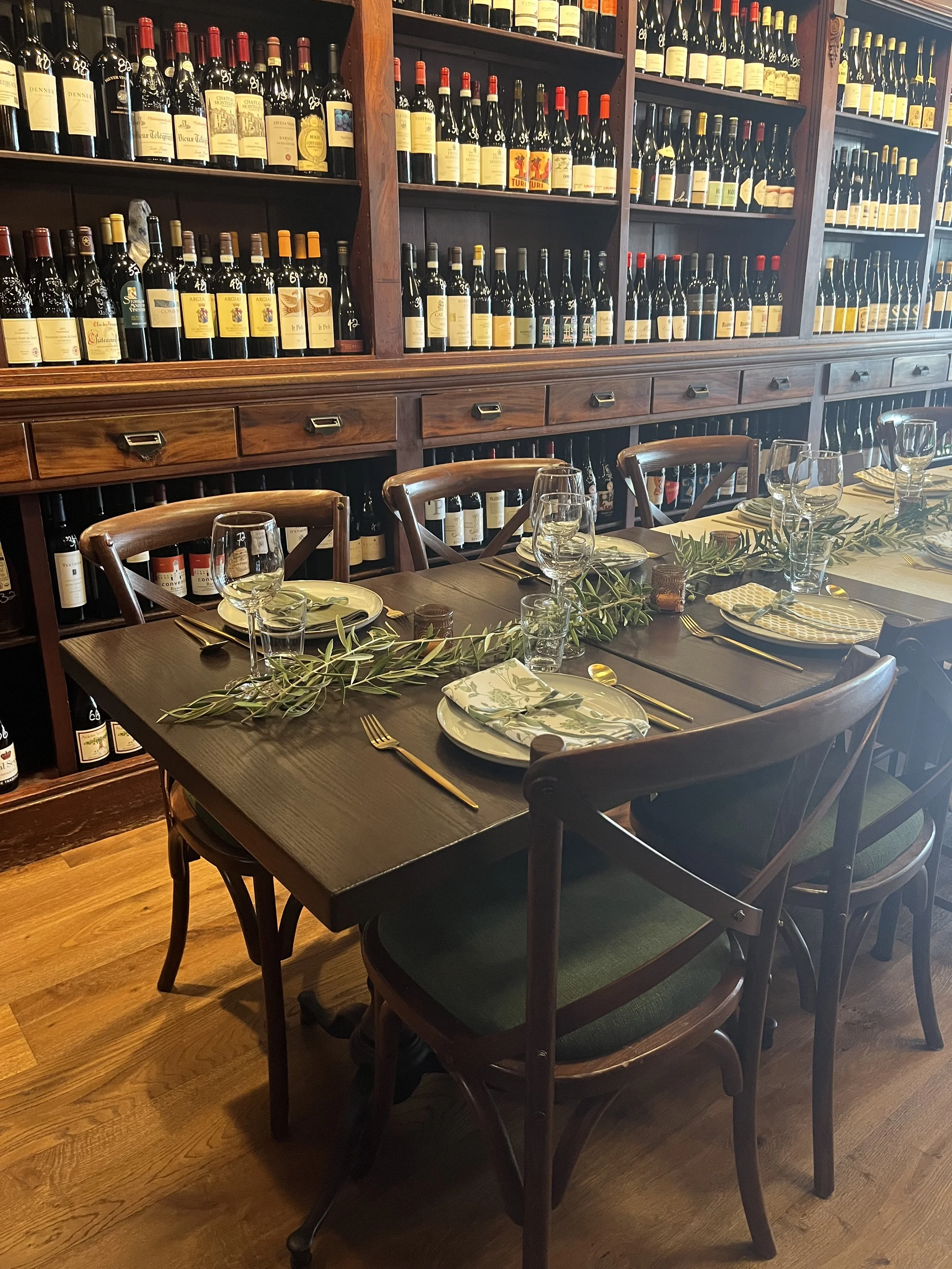 A dining table set with plates, glasses, cutlery, and napkins arranged for a meal, with a decorative greenery centerpiece. Behind the table, there is a large wooden wine rack filled with bottles of wine.