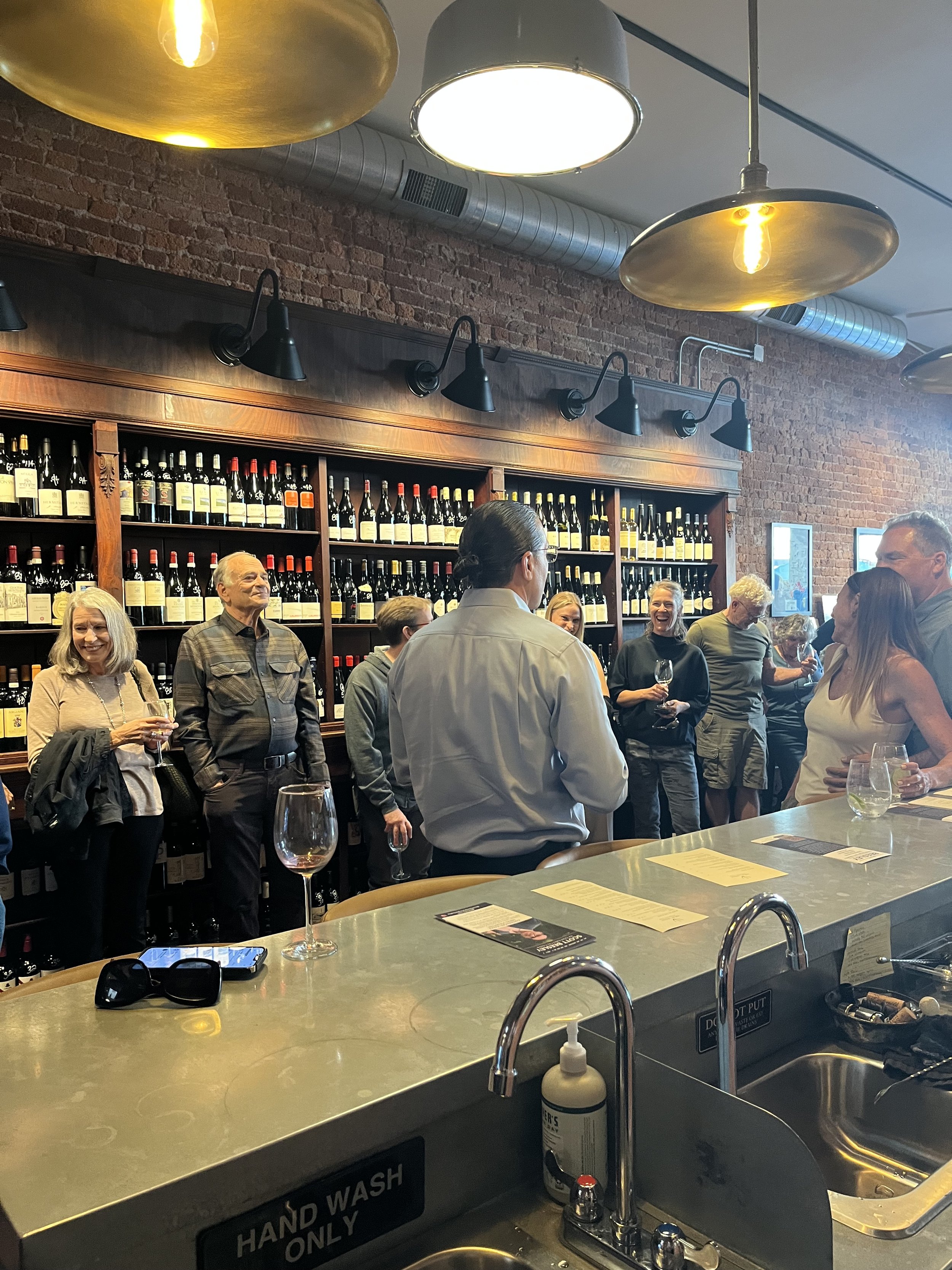 People socializing at a wine bar with shelves of wine bottles and industrial-style lighting.