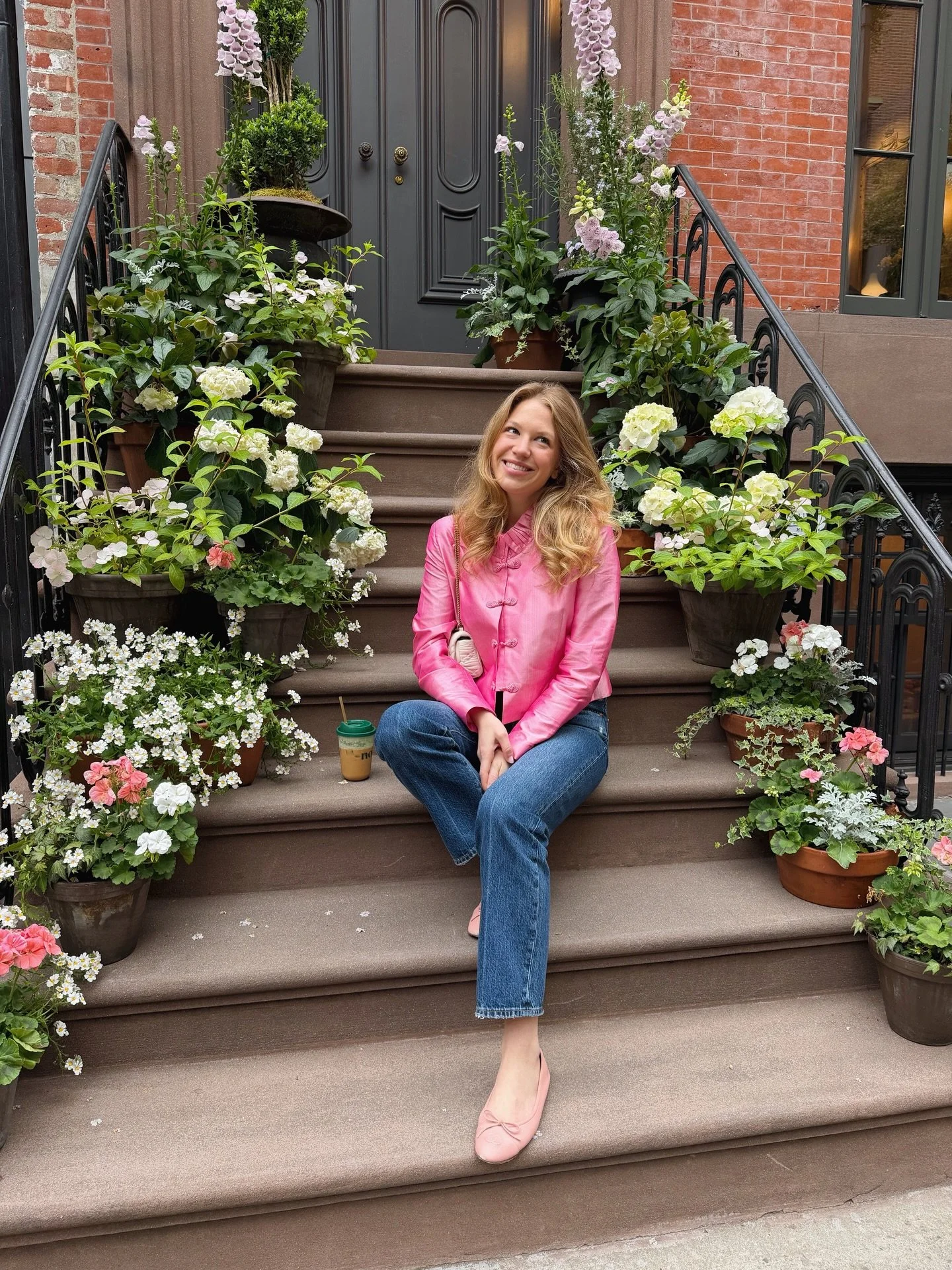 A woman in a pink jacket and blue jeans sitting on outdoor steps surrounded by potted flowers, smiling and looking to the side.