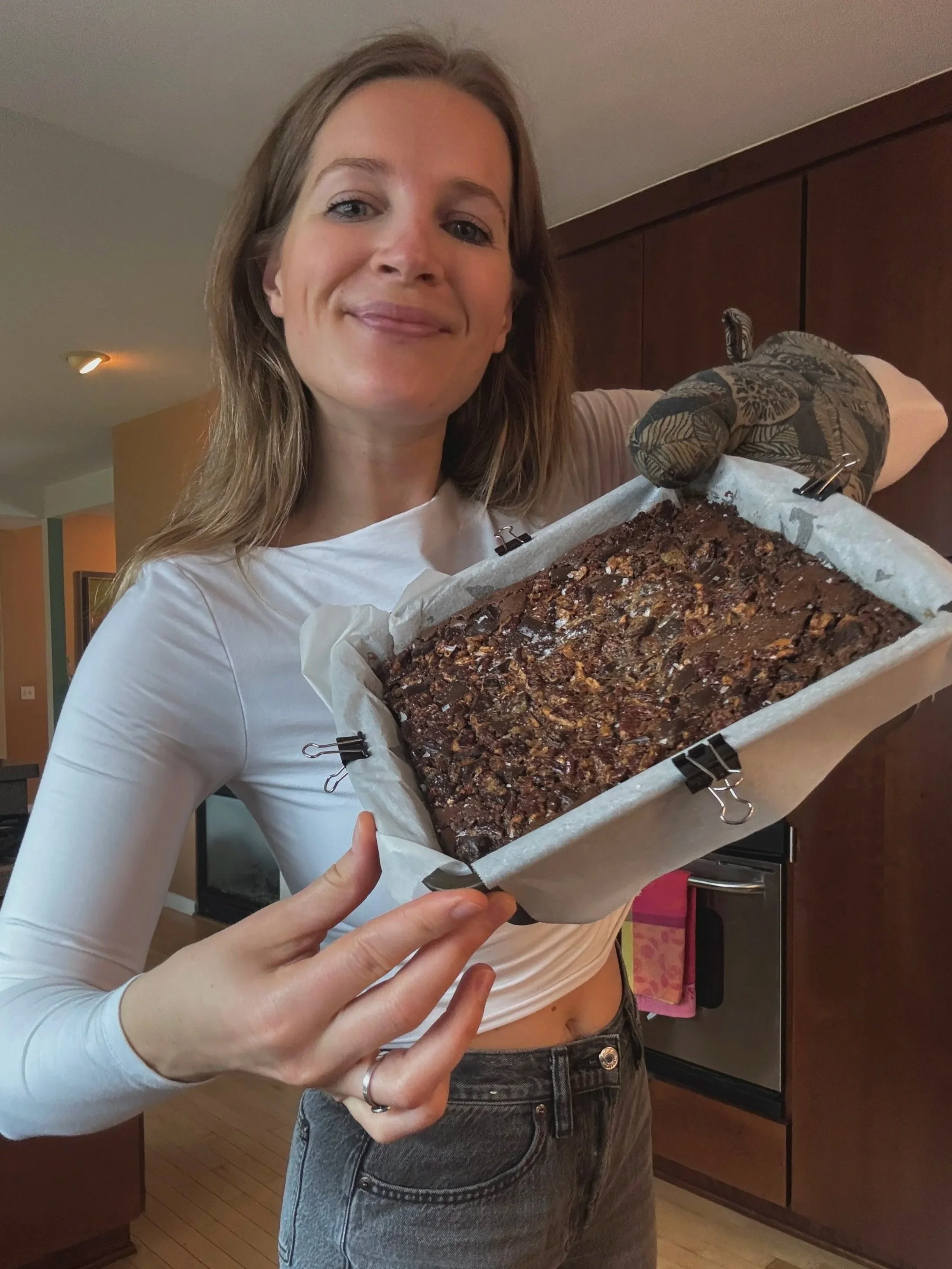 A woman with long blonde hair and a white long sleeve shirt is holding a freshly baked chocolate dessert with nuts on top inside a paper-lined baking tray.