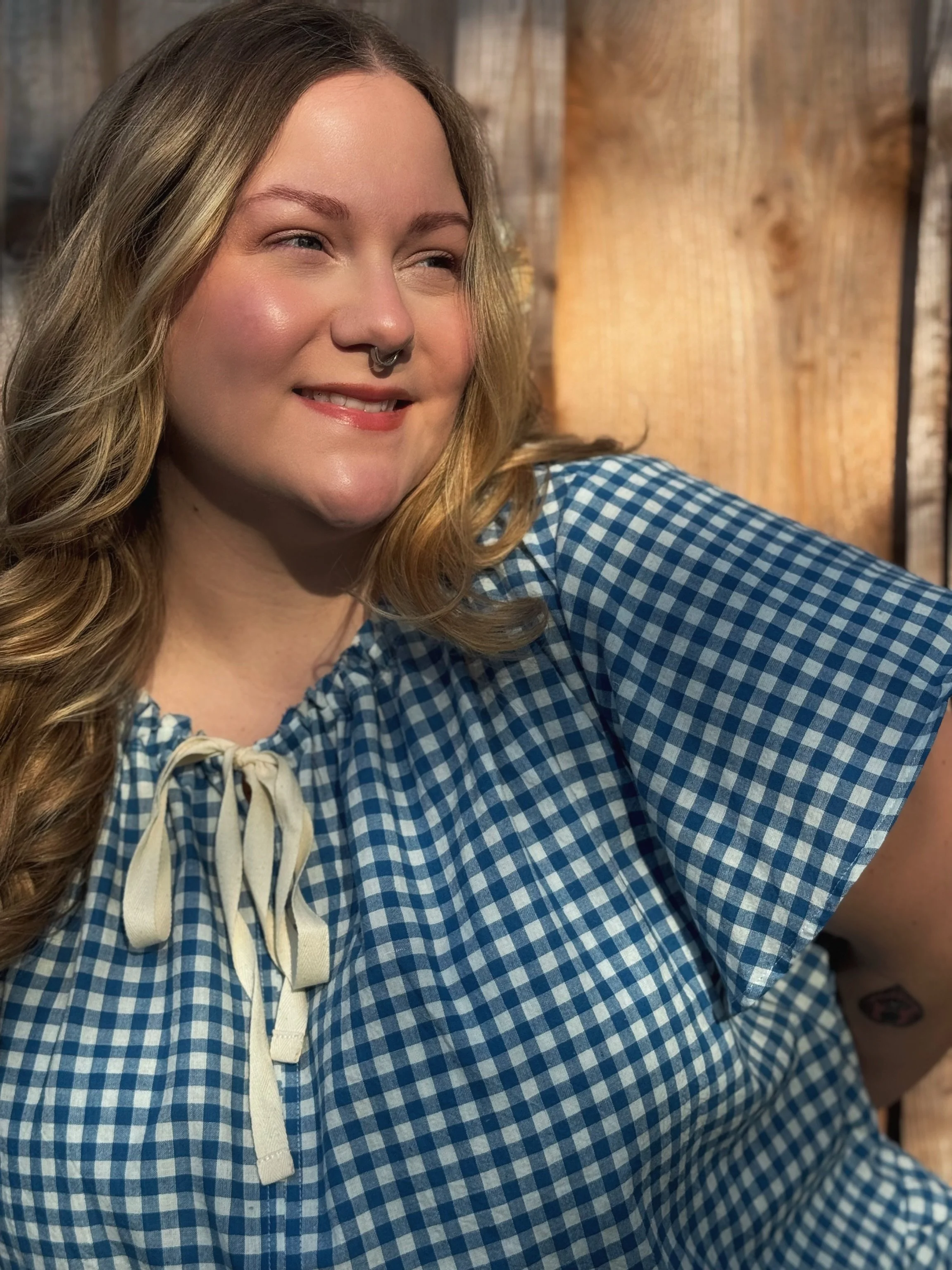 A woman with long wavy hair wearing a blue and white checkered shirt with a lace-up neckline, smiling while taking a selfie.