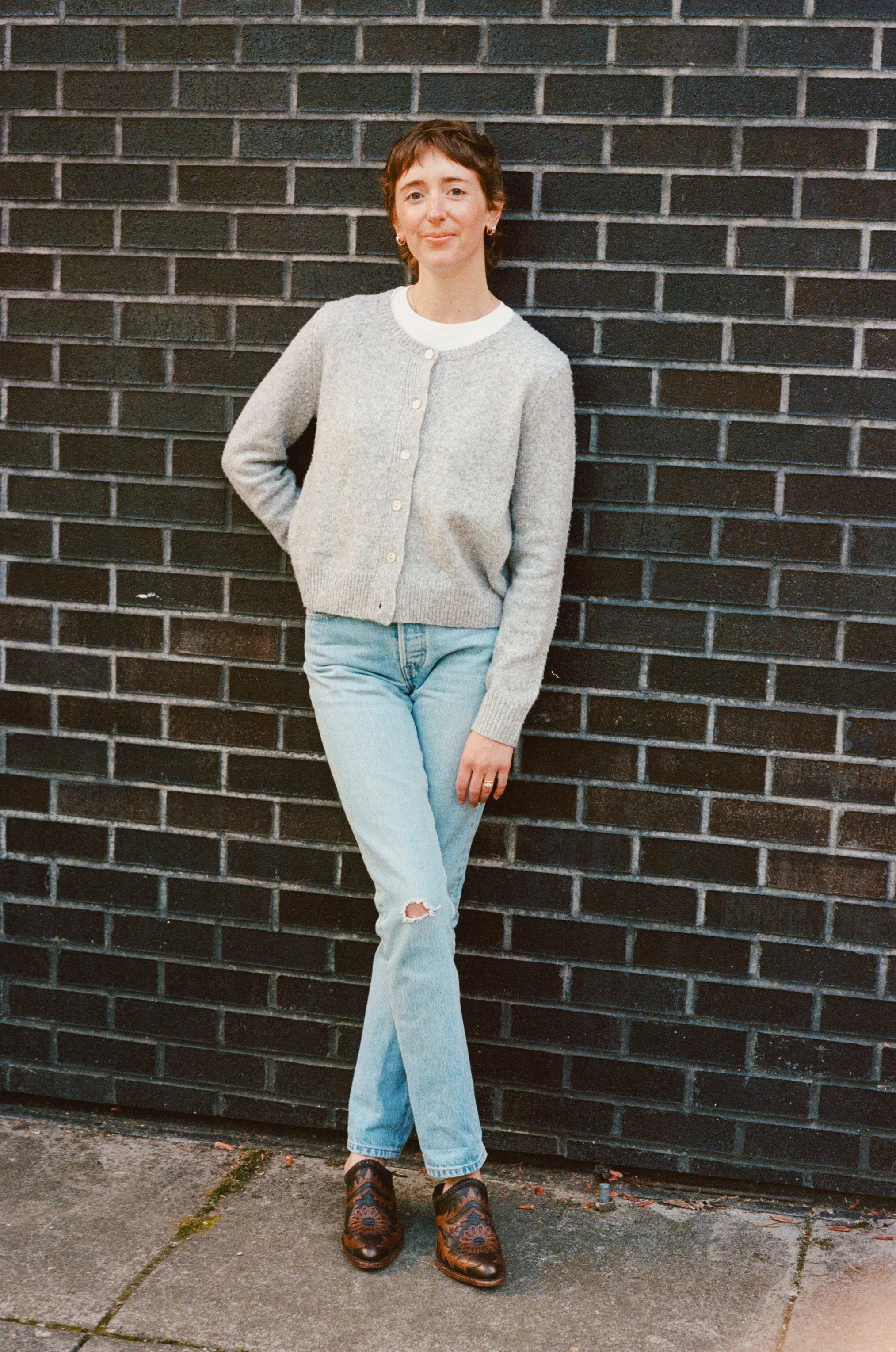 A woman with short hair standing against a black brick wall, wearing a gray cardigan, white shirt, blue jeans with a rip in the knee, and cowboy boots.