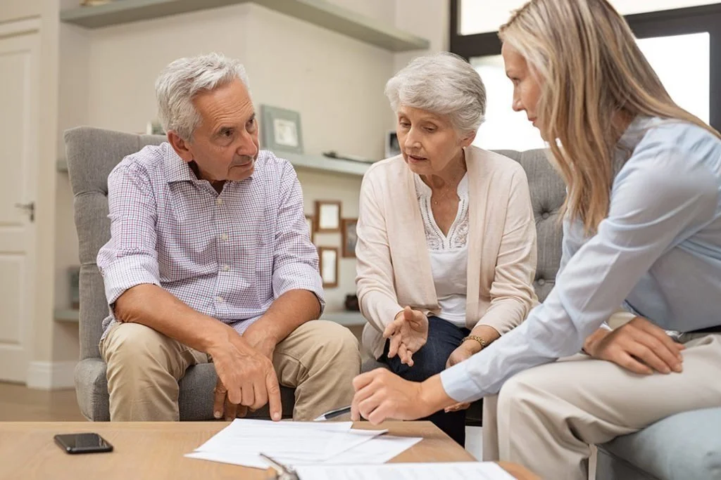 An elderly couple and a healthcare professional sitting together and reviewing documents in a living room.