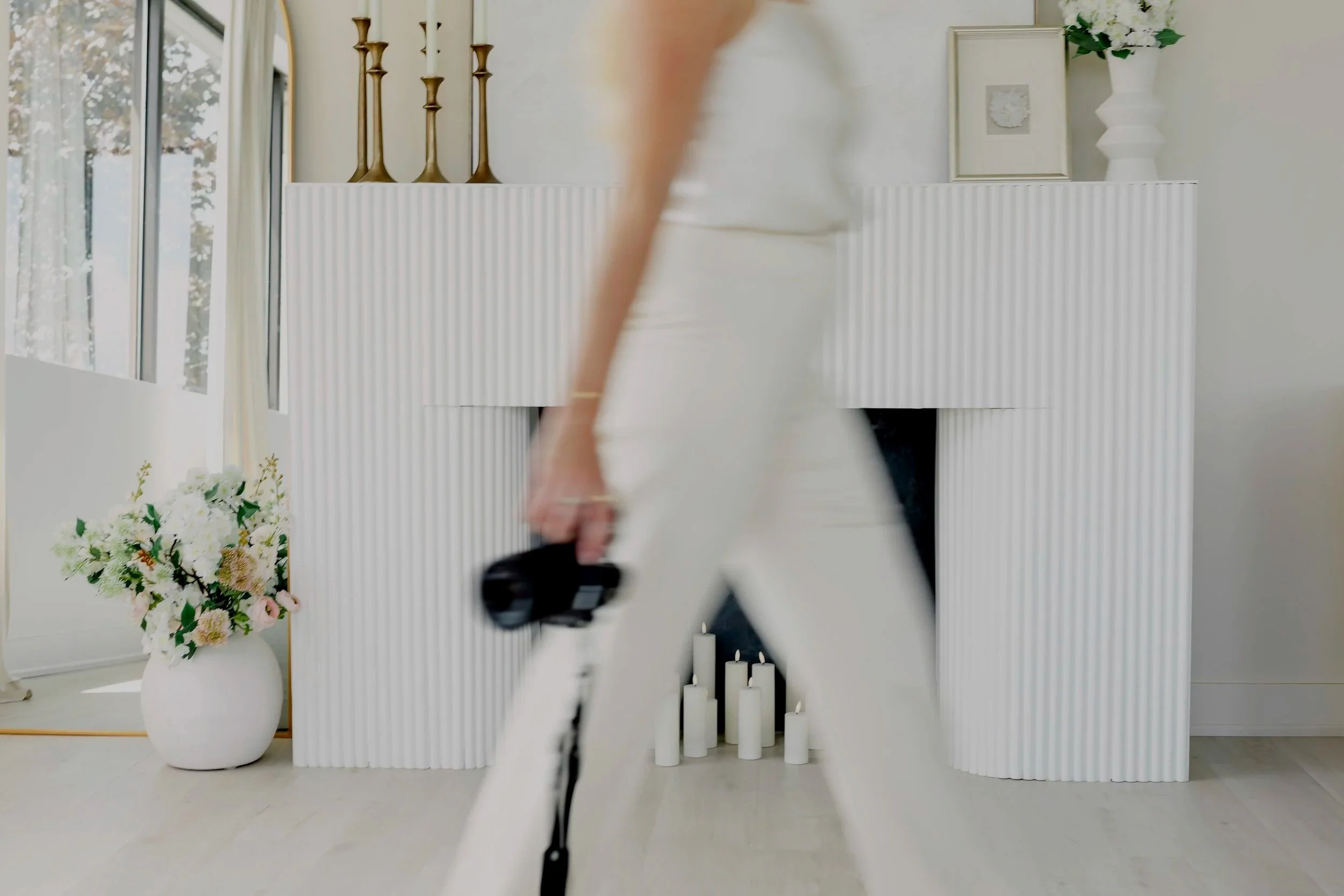 Person walking past a modern fireplace with white candles inside, a vase with flowers on the mantel, and decorative items including gold candlesticks and framed artwork in a bright living room.