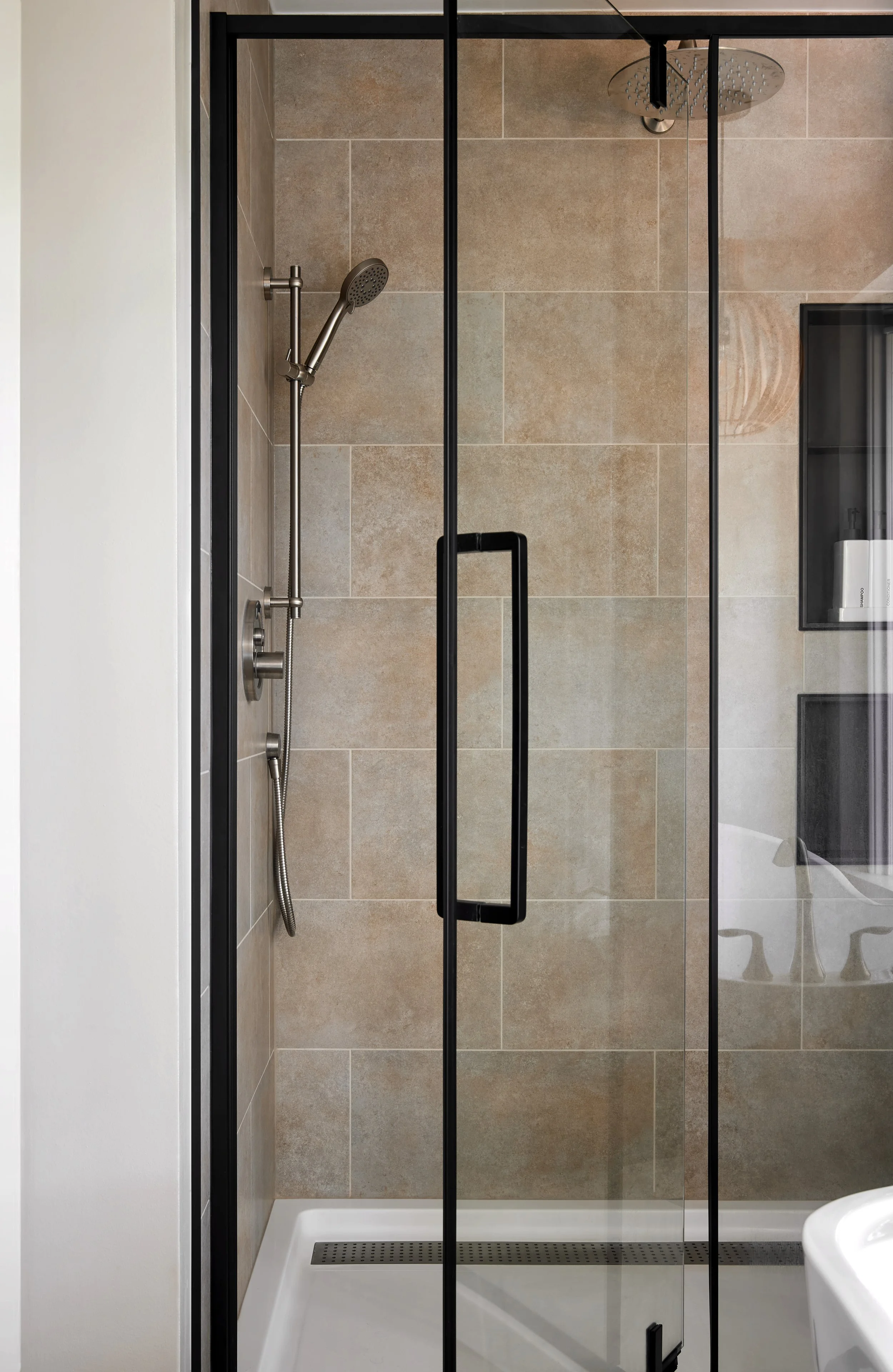 A modern walk-in shower with a glass door and black handle, beige tiles, a rainfall showerhead, a handheld shower, and a wall-mounted shelf.