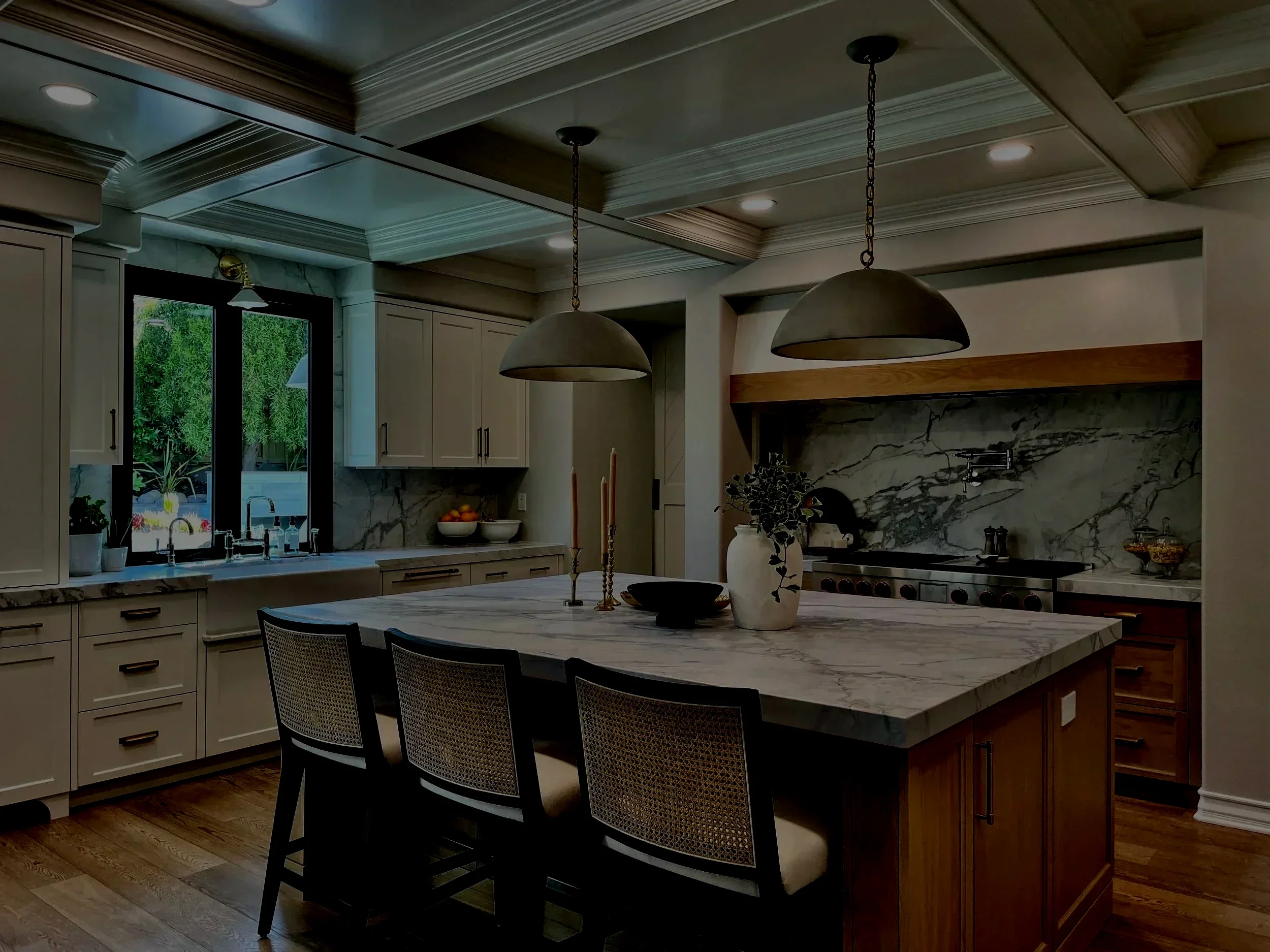 Modern kitchen with marble island, white cabinets, and black chairs. Two pendant lights hang over the island, and there is a window above the sink showing outdoor greenery. The back wall has a marble backsplash and a stove.