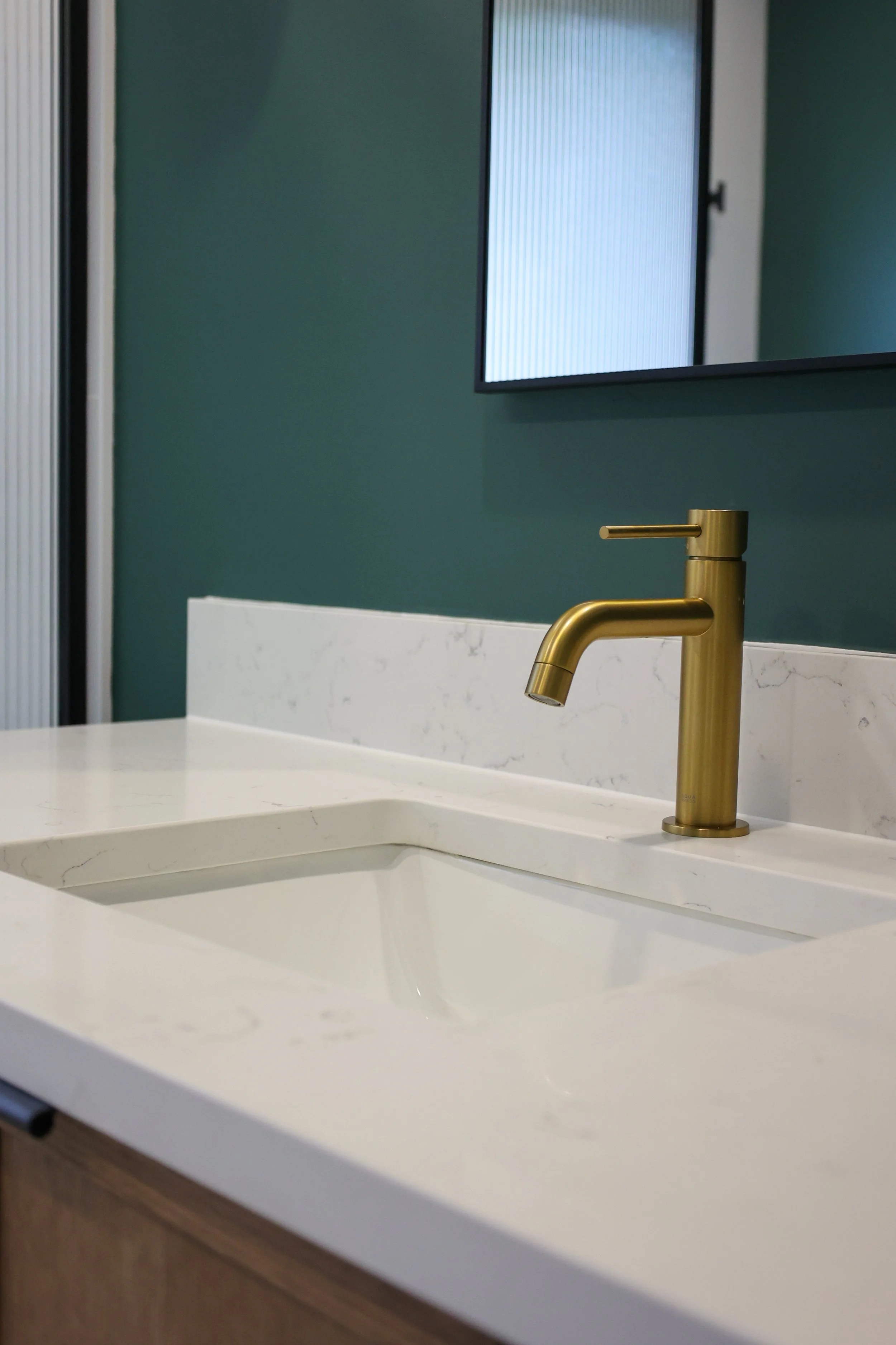 Close-up of a modern bathroom sink with a gold faucet on a white marble countertop, against a green wall with a mounted TV.
