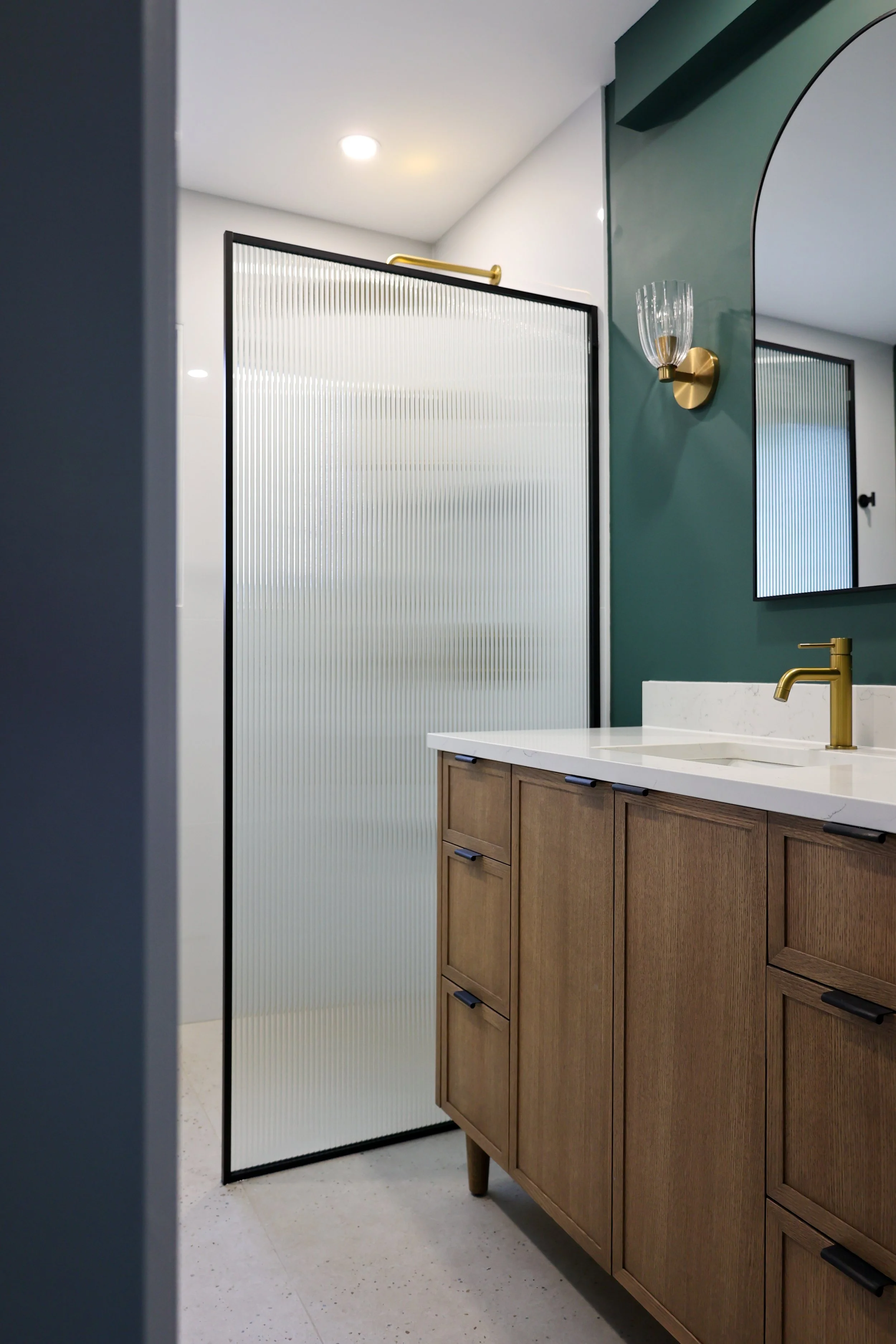 Modern bathroom with a wooden vanity, white countertop, gold faucet, and a textured glass shower door.