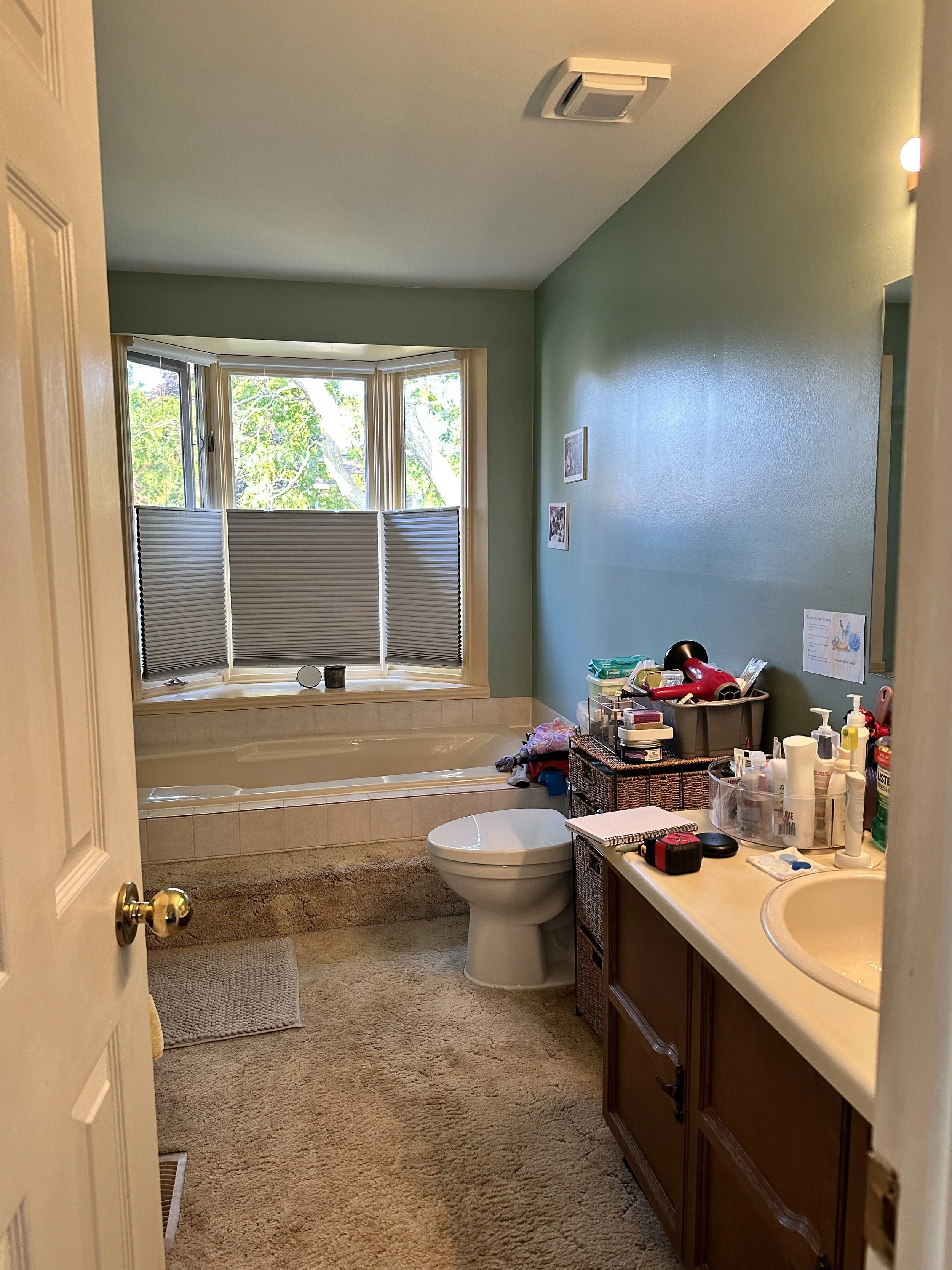 A cluttered bathroom with a window, bathtub, toilet, bathroom sink, and various toiletries and items on the counter.
