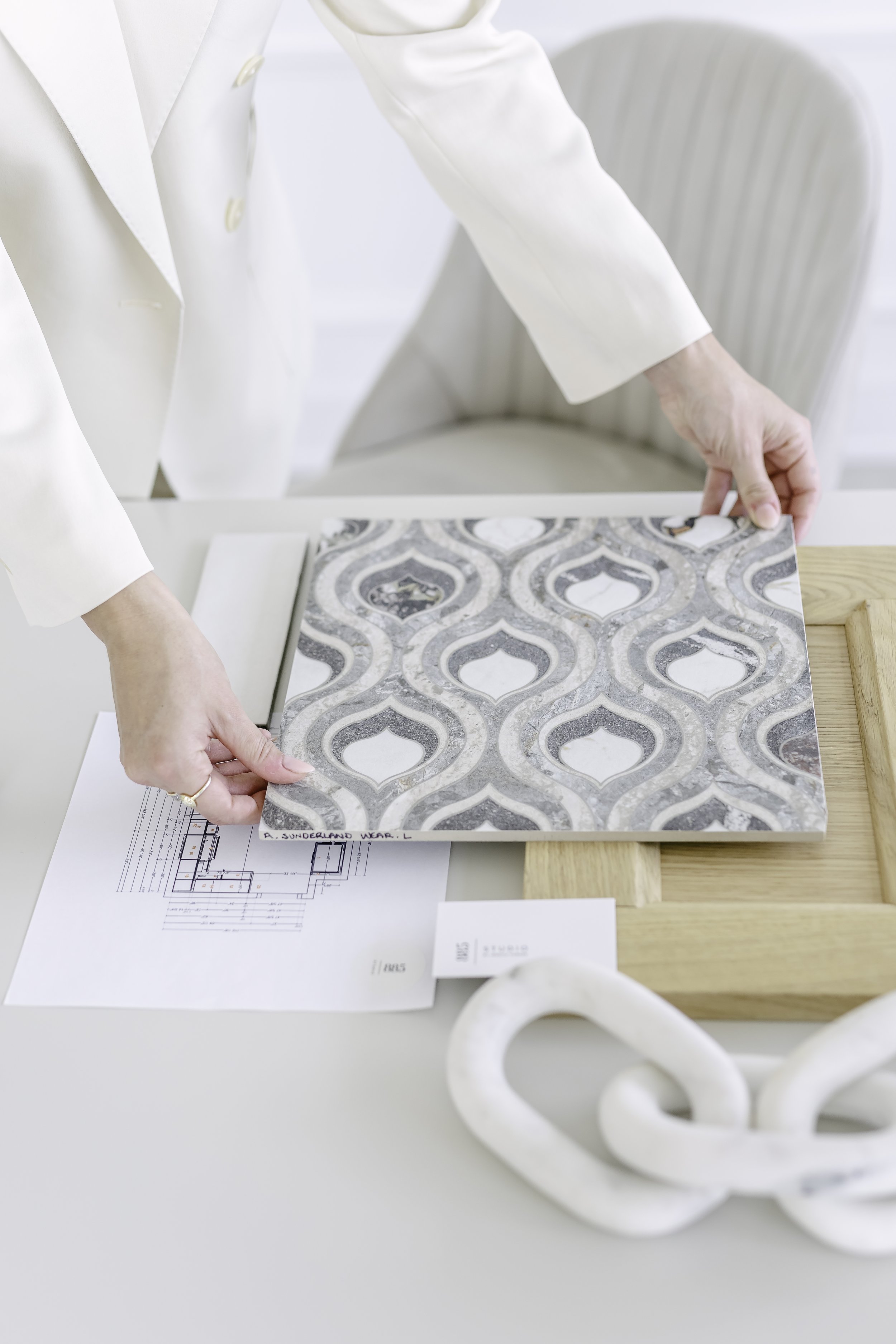 Person in white blazer examining a tile sample with a patterned design, on a white desk with architectural plans and decorative objects.