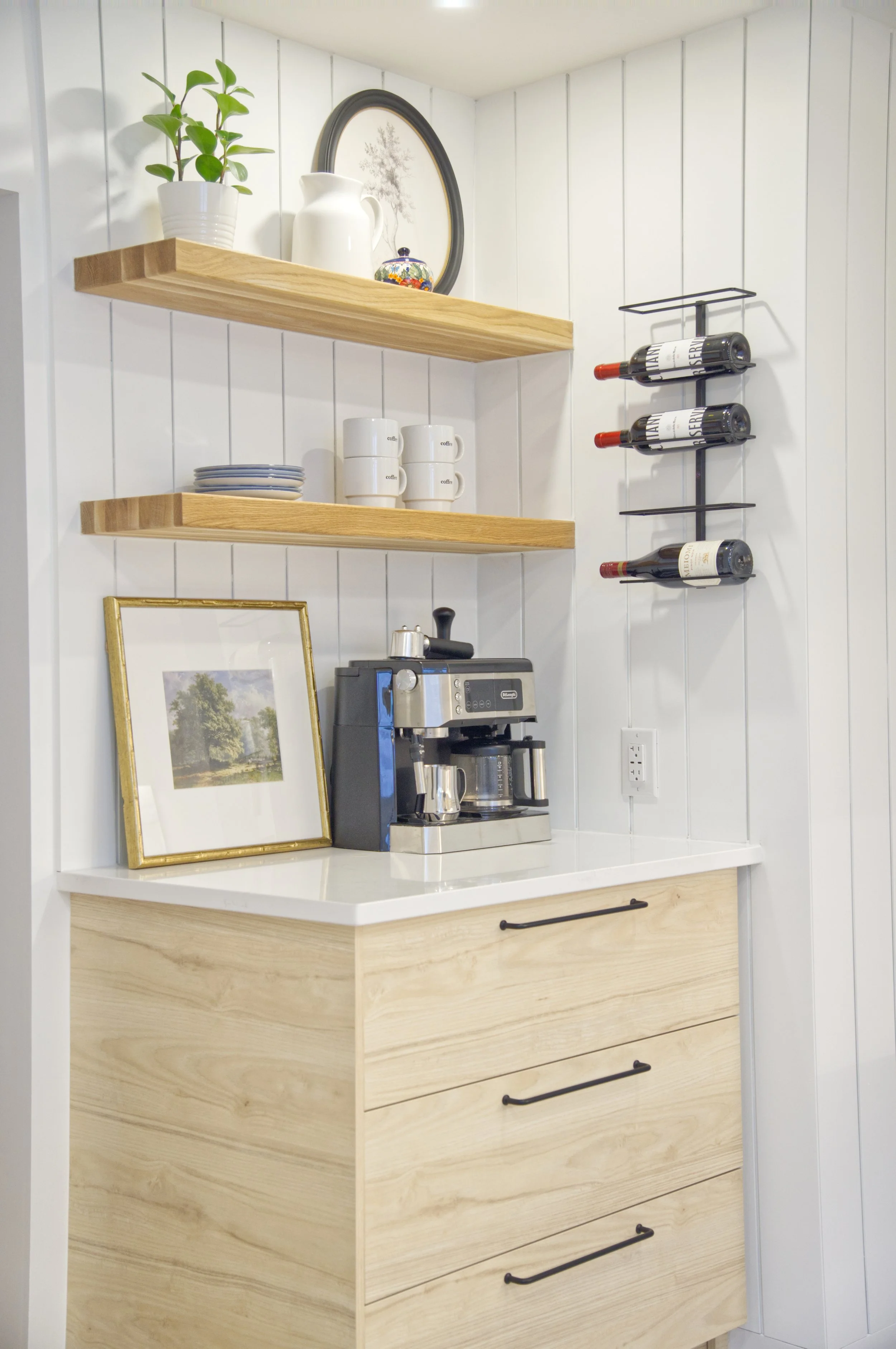 This image depicts a cozy kitchen corner with white paneled walls, wooden shelves, a coffee maker, a framed picture, and a wine rack with bottles.