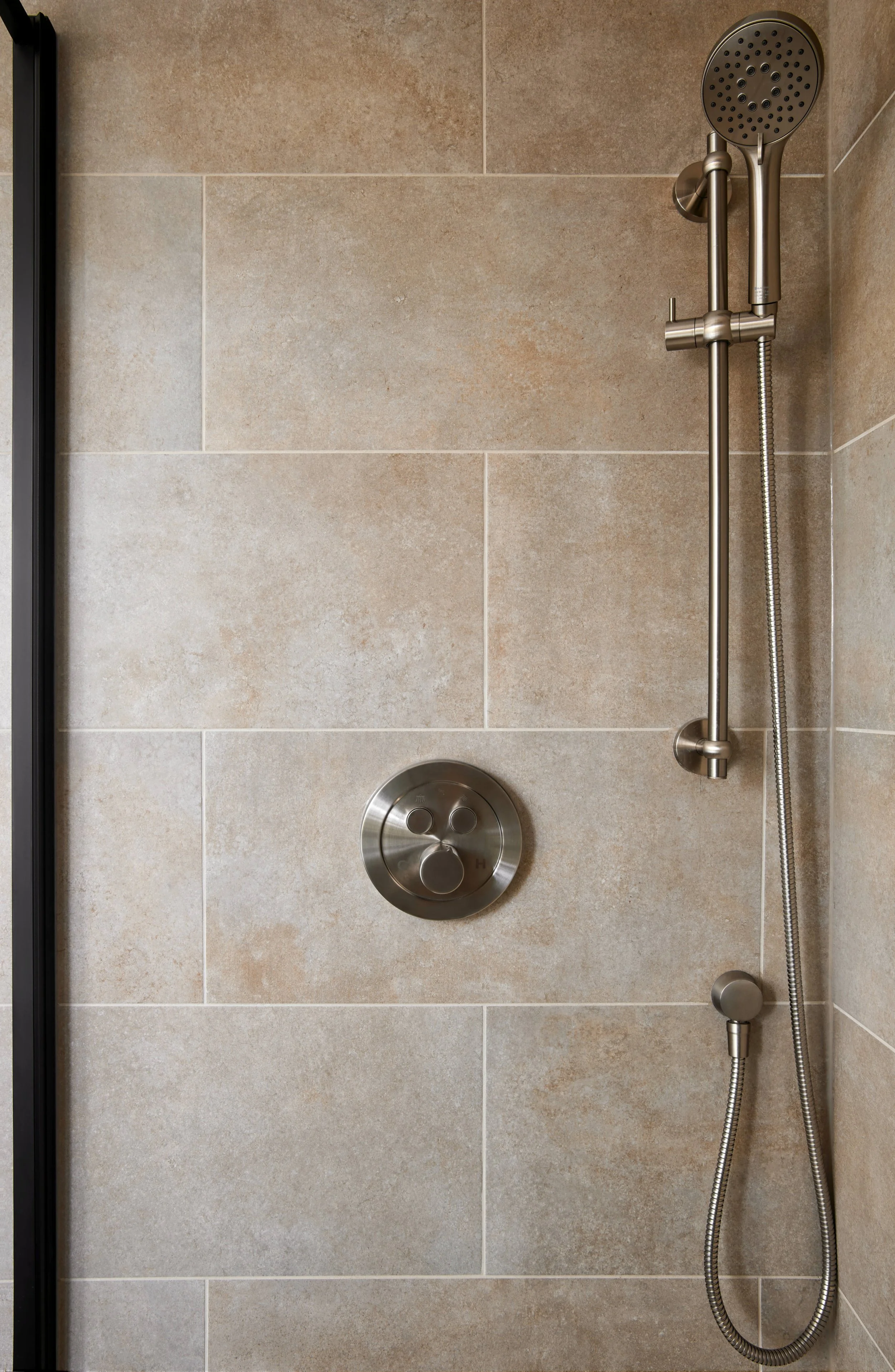 A modern shower with beige tile walls, a silver adjustable showerhead, a handheld showerhead, and a circular temperature control knob.