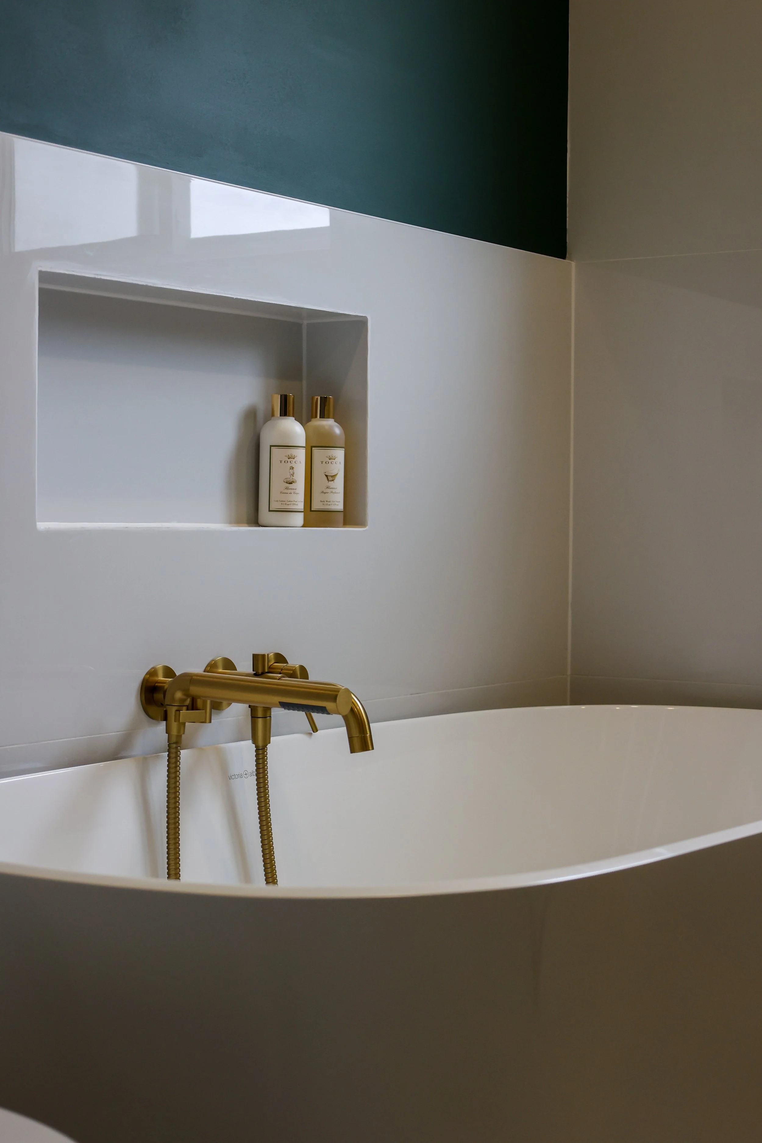 Bathroom with a white bathtub, gold faucet, a built-in shelf with two bottles of toiletries, and a two-tone wall in teal and beige.