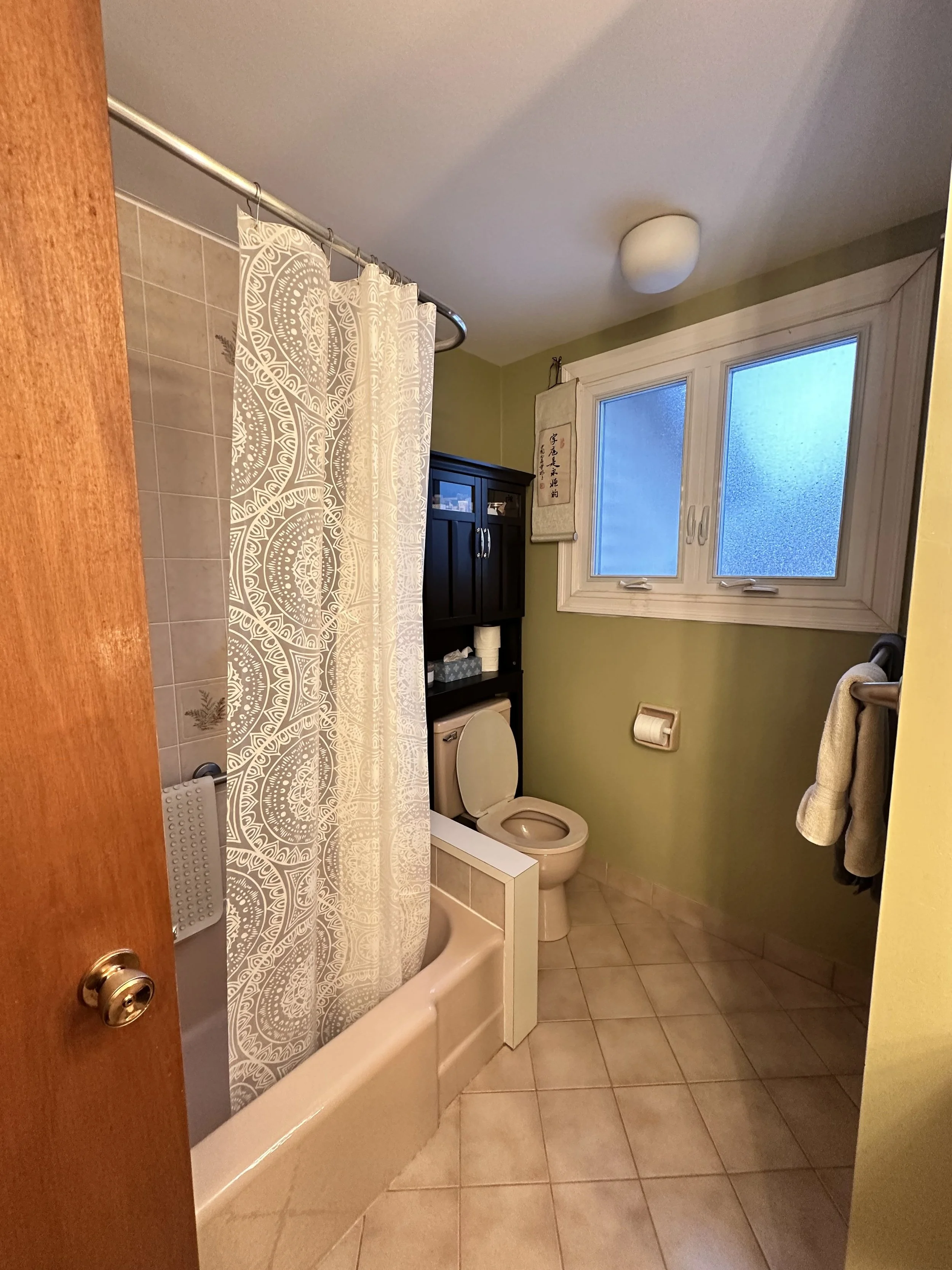 A small bathroom with a green wall and beige tiled floor. There is a bathtub with a patterned shower curtain, a toilet with a black shelf above it, and a window with frosted glass. Towel rack with towels and a wall hanging with Asian characters.
