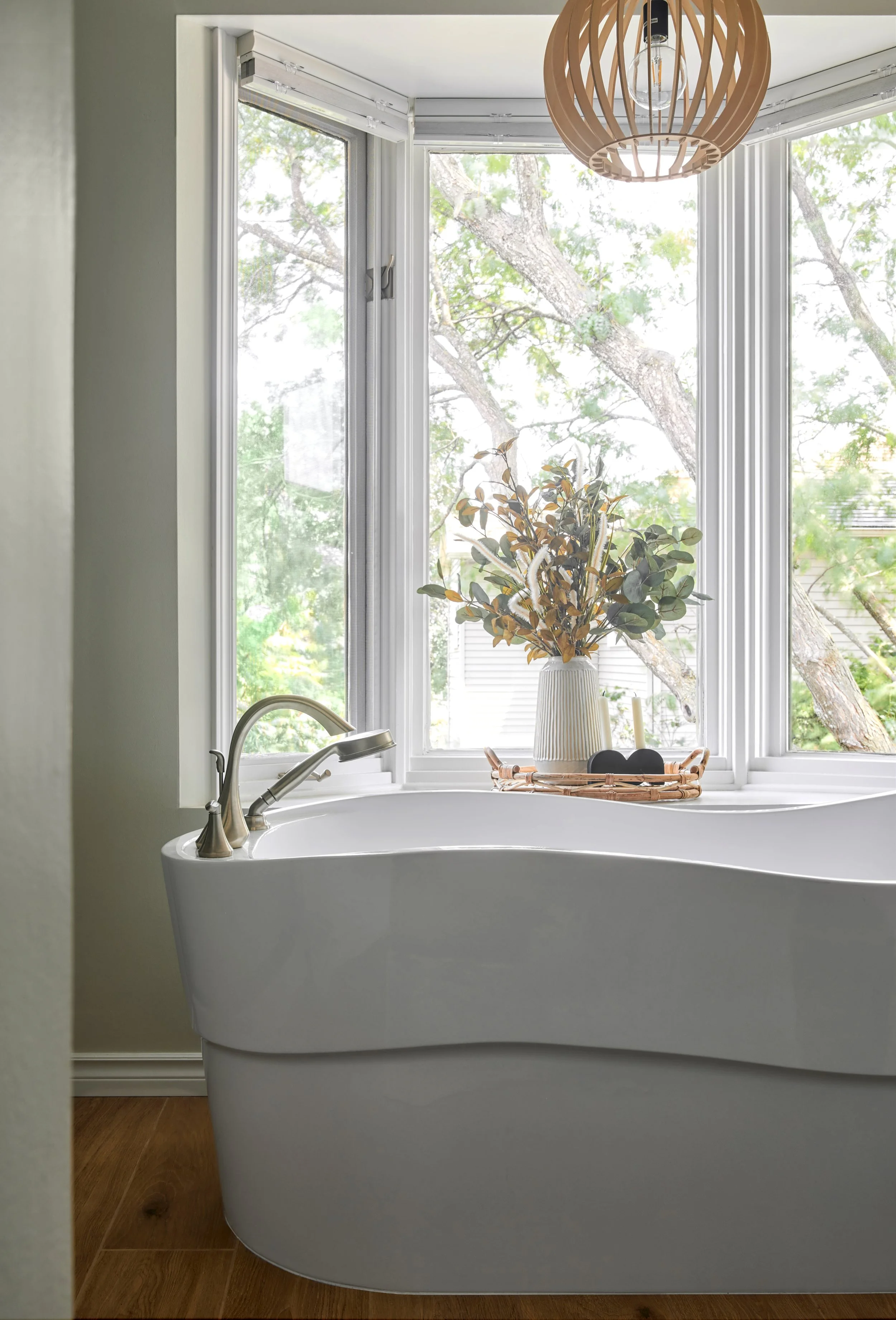 White freestanding bathtub in front of a bay window with a vase of dried flowers and a tray holding bathing essentials, with a wooden pendant light hanging from the ceiling.