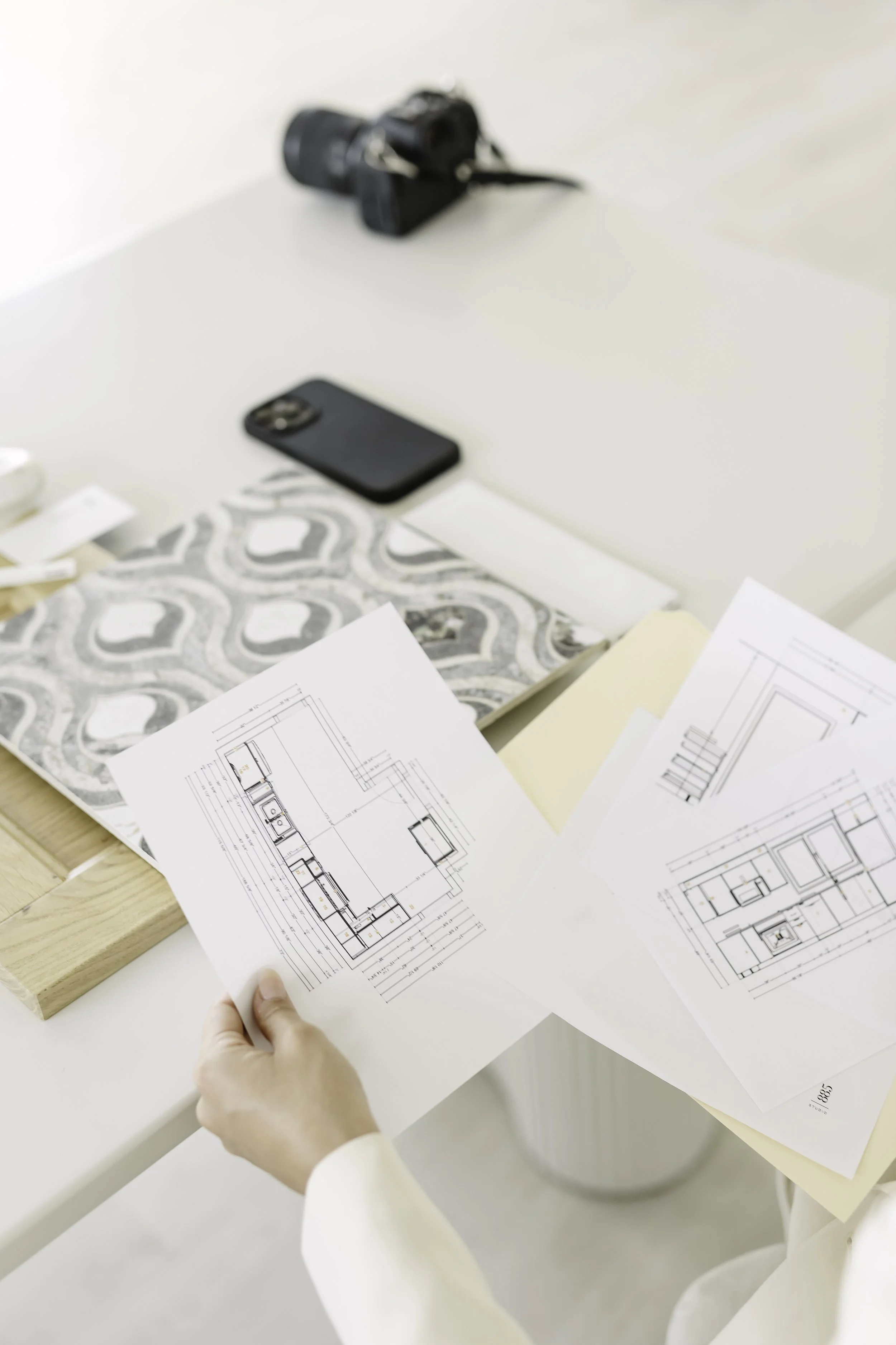 Person reviewing architectural blueprints on a desk, with a smartphone, camera, and swatch sample nearby.