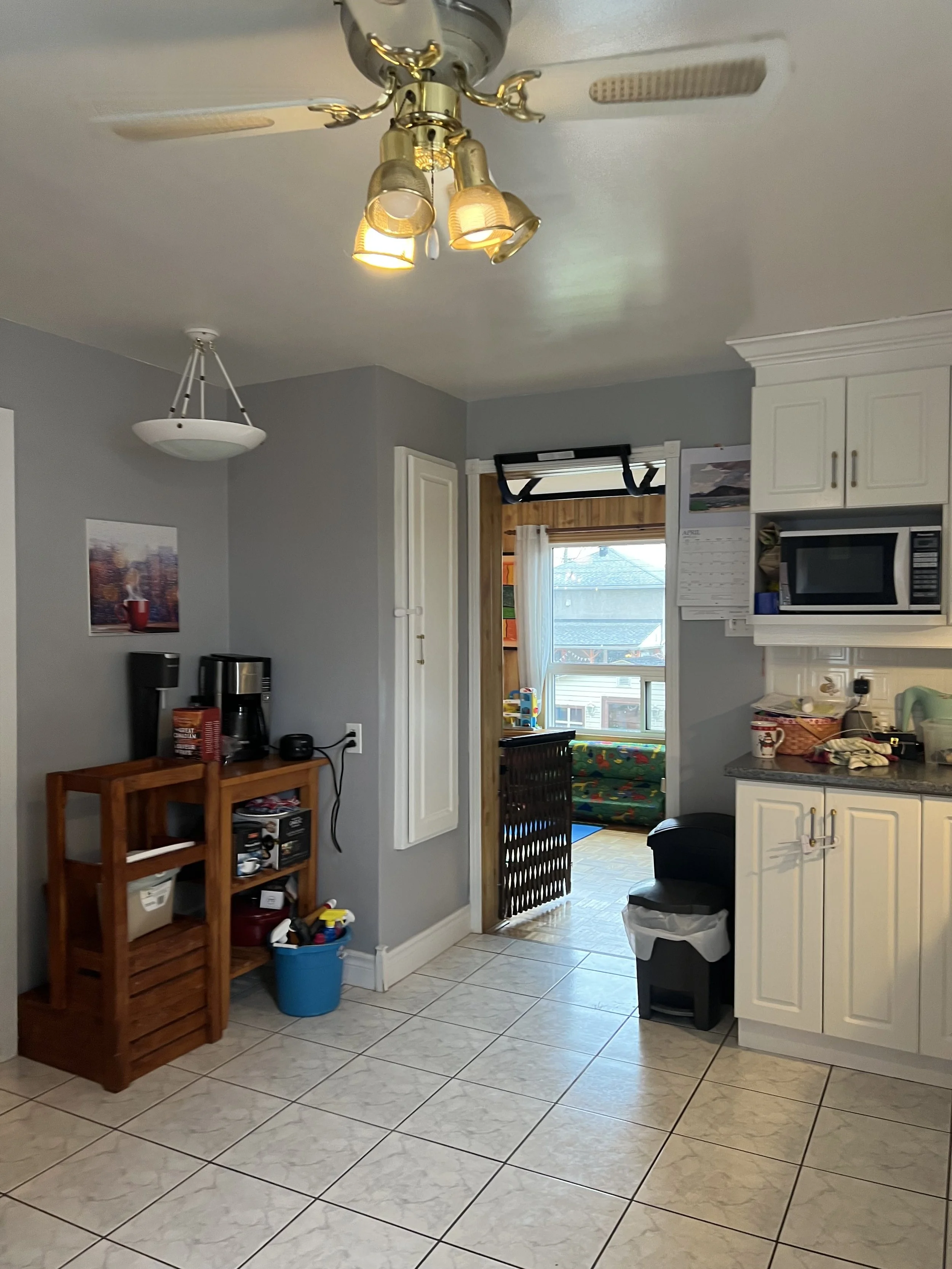 A kitchen with gray walls, white cabinets, tile flooring, and a wooden doorway leading to a sunlit room with a window and a colorful couch. There is a ceiling fan with lights, a wooden shelving unit with small appliances, and various items on the cou