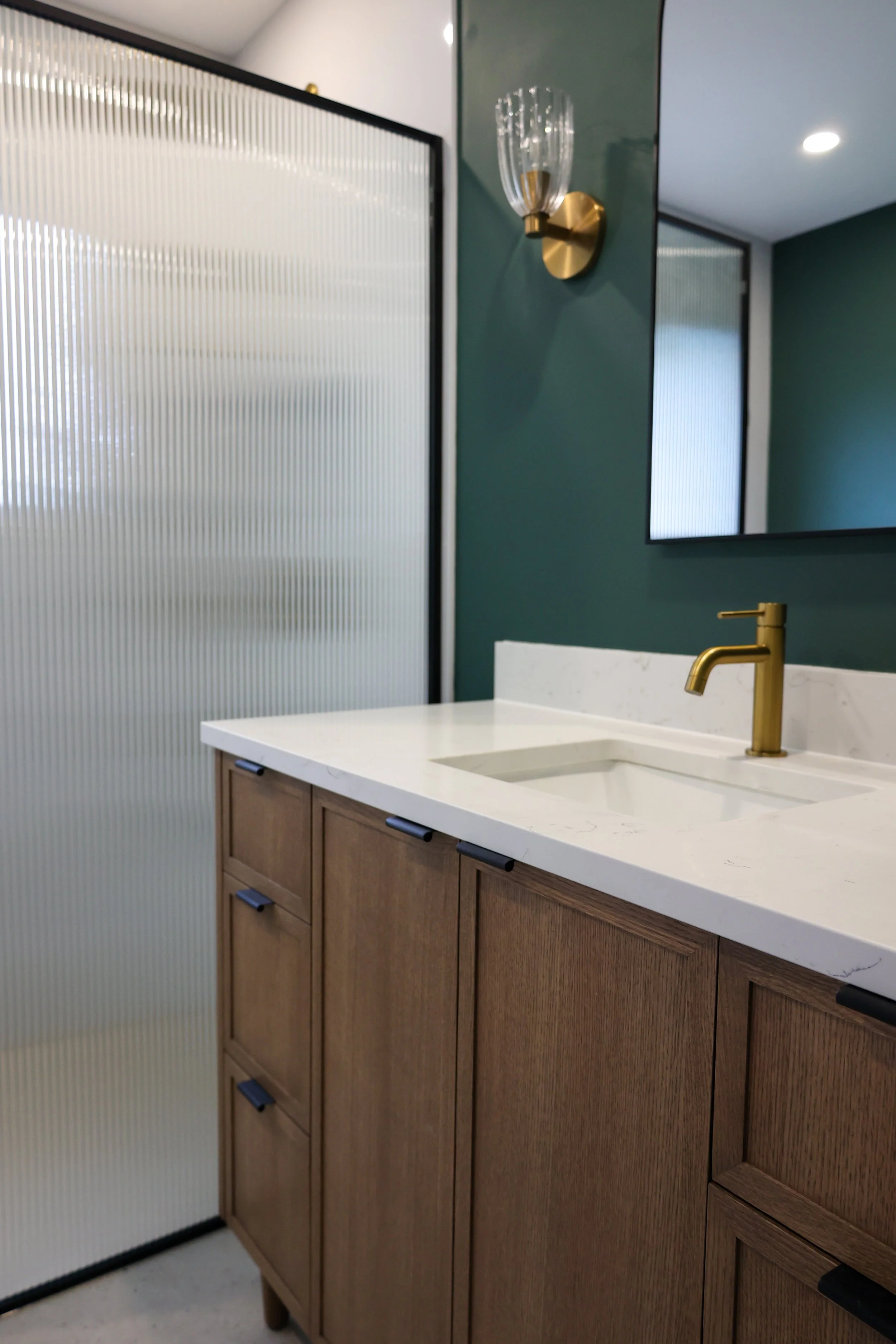 Modern bathroom with wooden cabinet, white marble countertop, gold faucet, textured shower enclosure, and teal wall with a gold wall light.