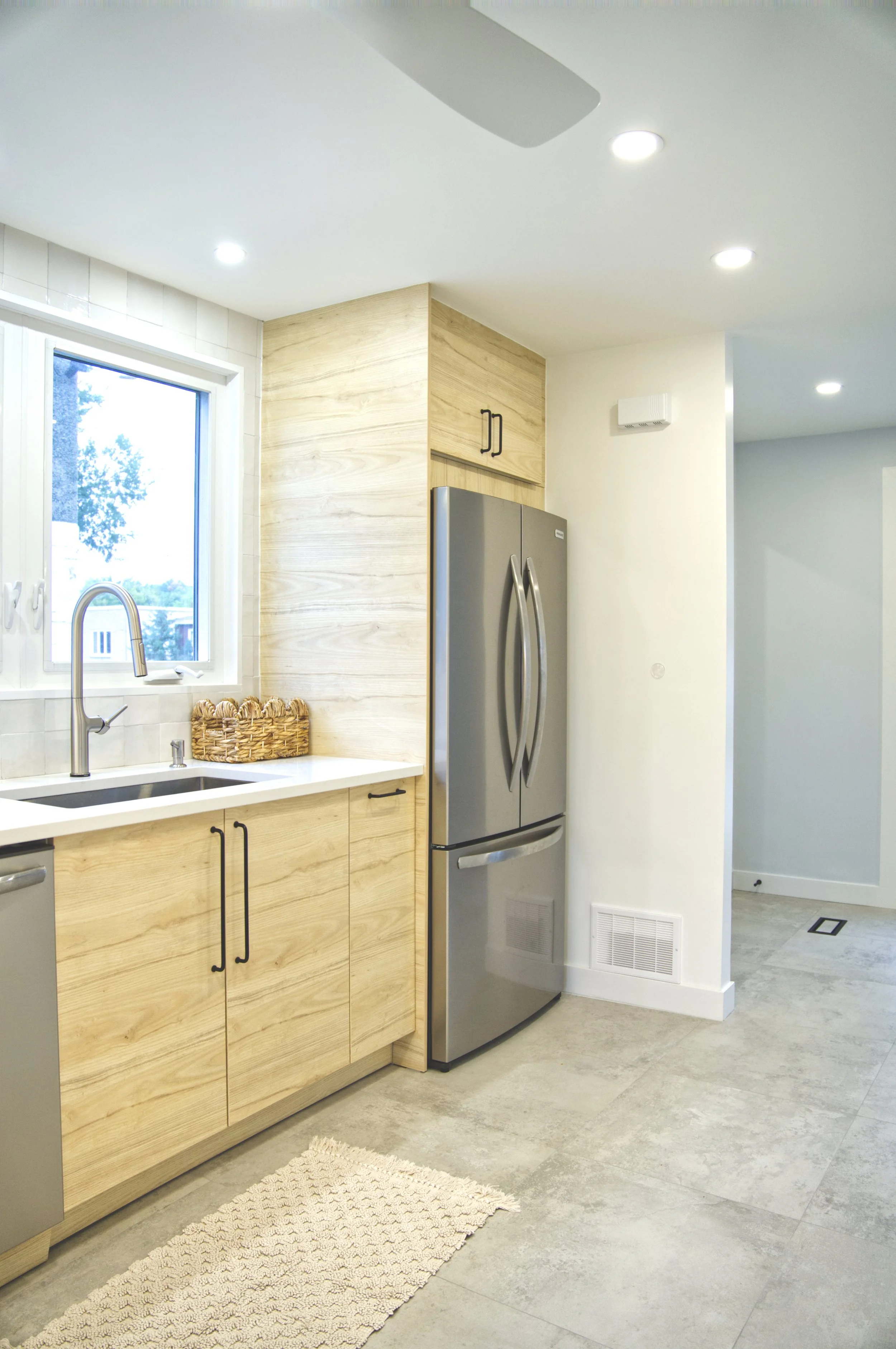 Modern kitchen with natural wood cabinets, stainless steel refrigerator, white countertop, and a window above the sink, with neutral-colored tiles and a small woven rug.