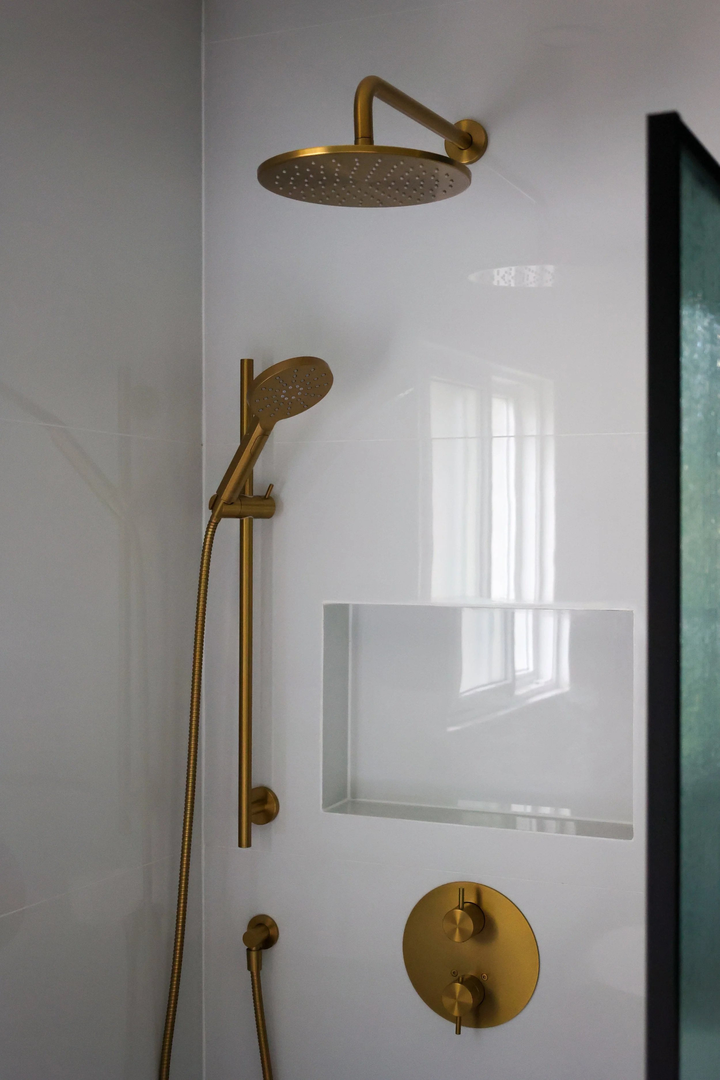 Modern shower with gold fixtures, a large overhead rain showerhead, a handheld showerhead, a built-in niche, and a round control panel, all mounted on white tiled wall.