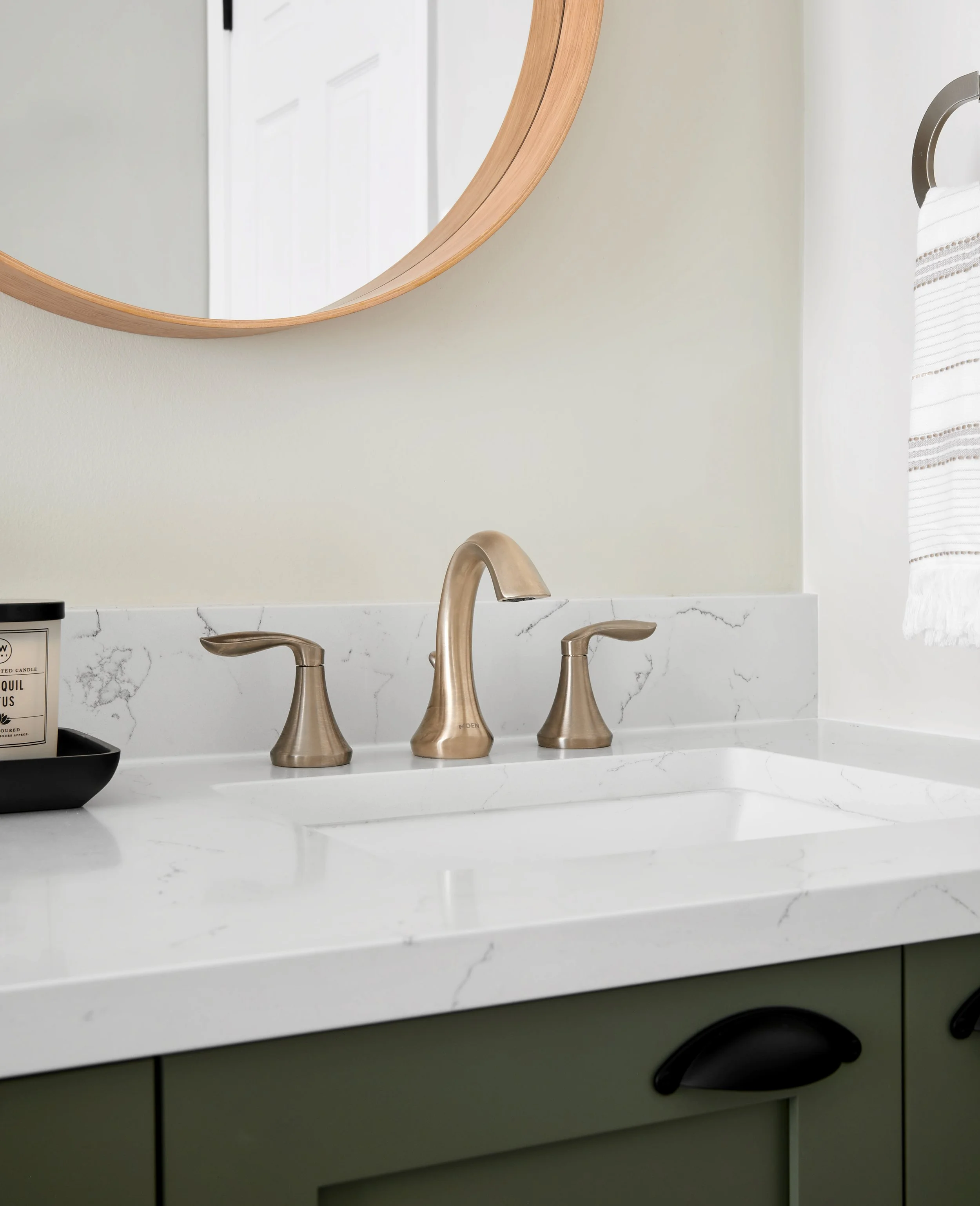 Bathroom sink with a white marble countertop, brass faucet, and two matching brass handles. A mirror with a wooden frame hangs above the sink, and a towel is on a ring to the right. A black tray with a candle is on the left side of the sink.