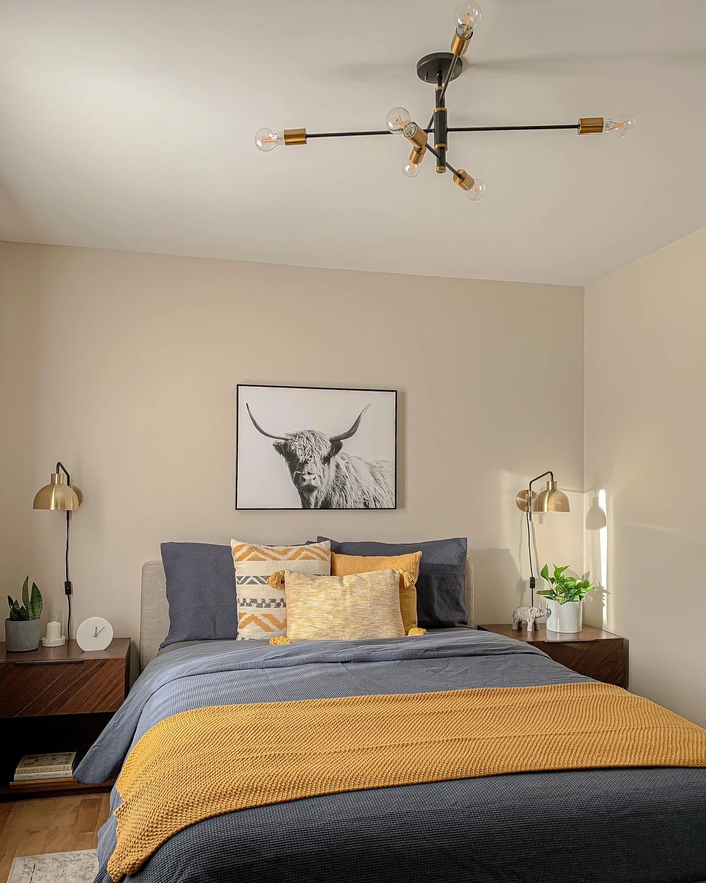 A bedroom with a bed featuring dark and yellow pillows, a gray blanket, and a yellow throw blanket. Two wooden nightstands with potted plants, lamps, and decorative items. A black and white framed photo of a highland cow hangs above the bed. A modern chandelier with exposed bulbs is on the ceiling.