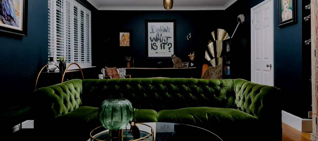 Living room with dark blue walls, white window shutters, a green velvet tufted sofa, artwork on the walls, and a black framed mirror or artwork with the phrase 'I want in what is it?' in the center of the back wall.