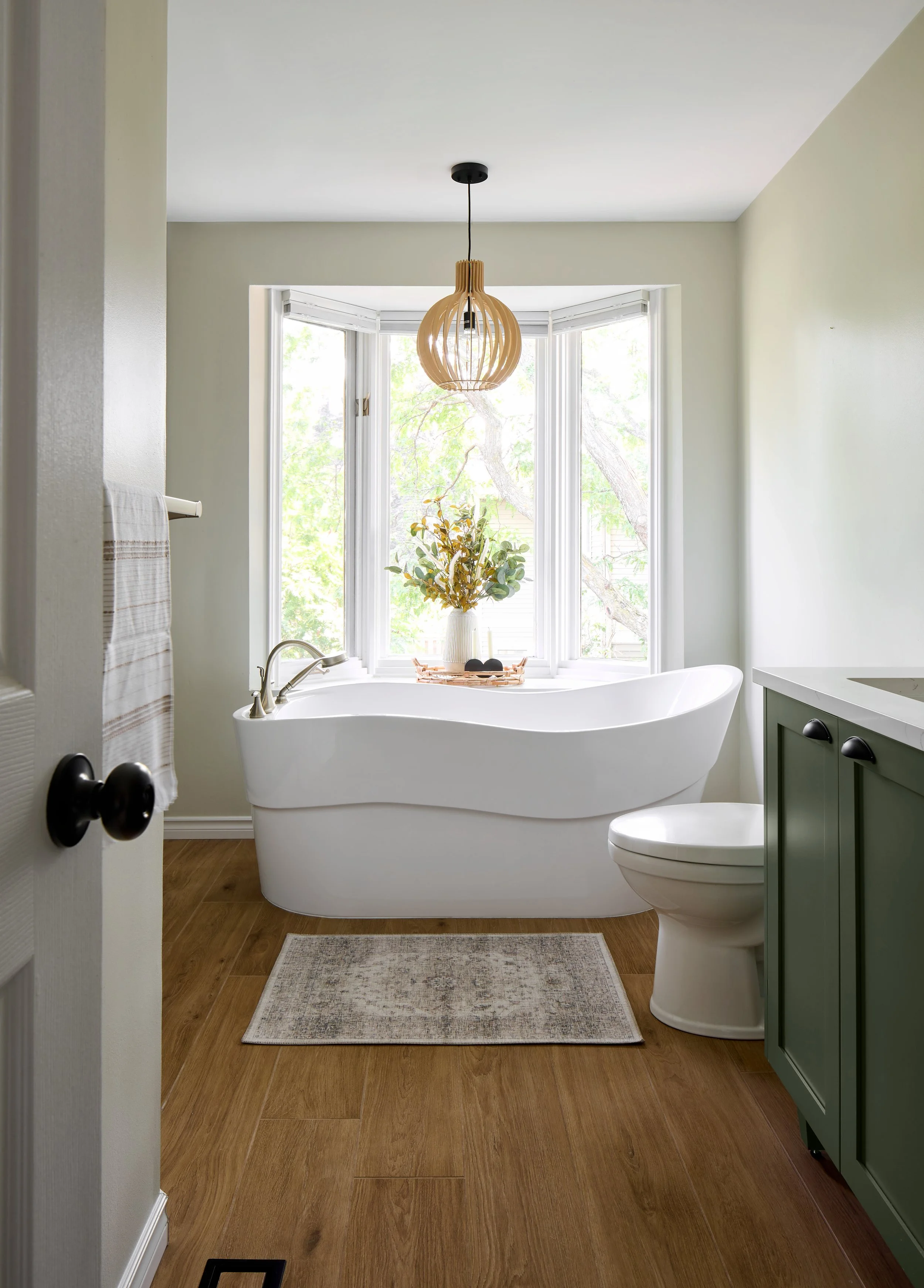 Bright bathroom with a free-standing bathtub in front of a large bay window, a green vanity cabinet, and a towel on a wall rack.