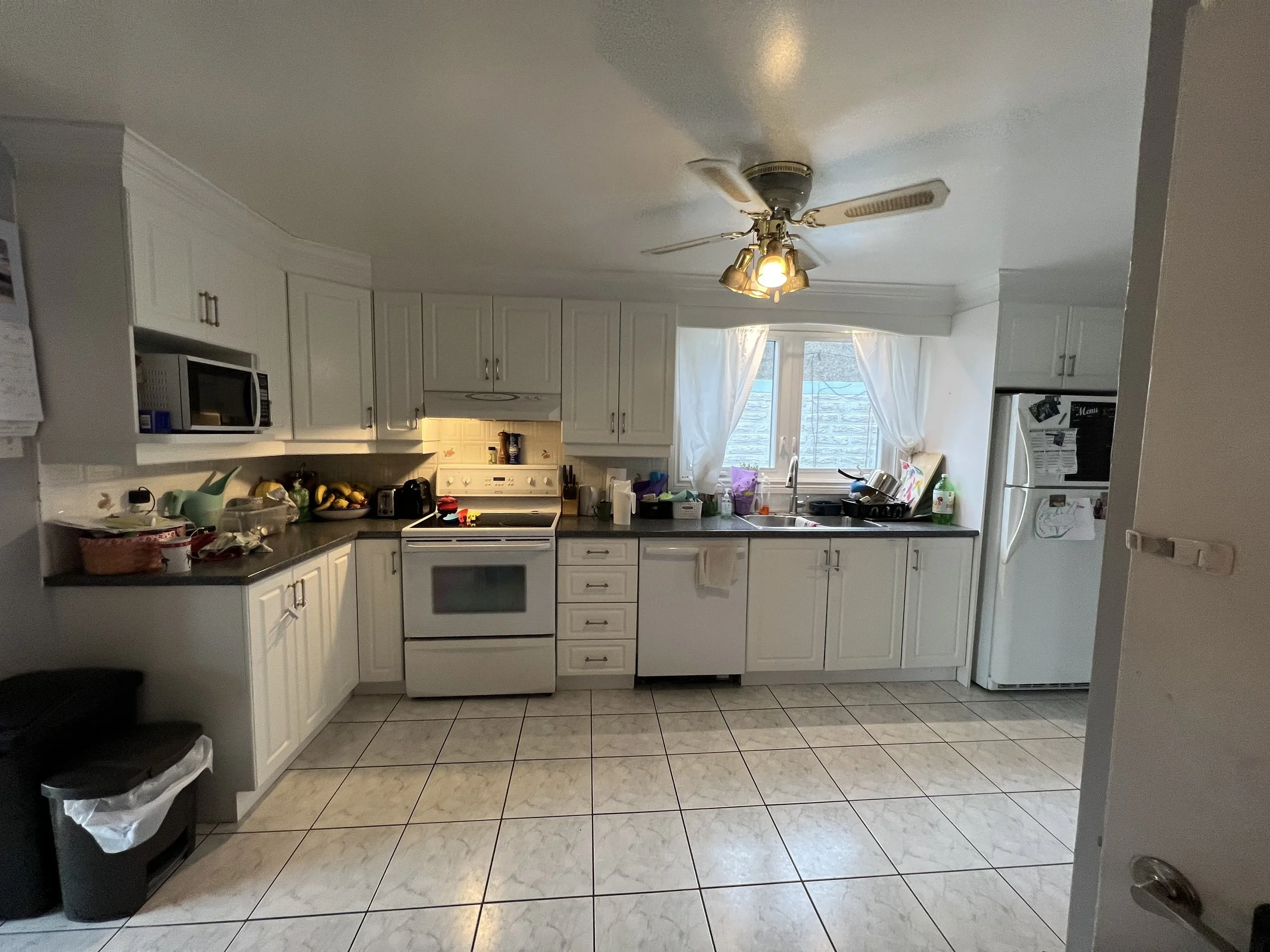 Kitchen with white cabinets, black countertops, and a white stove. There are various items on the counters, including fruit, dishes, and containers. A window with white curtains lets in natural light. A refrigerator is on the right side.