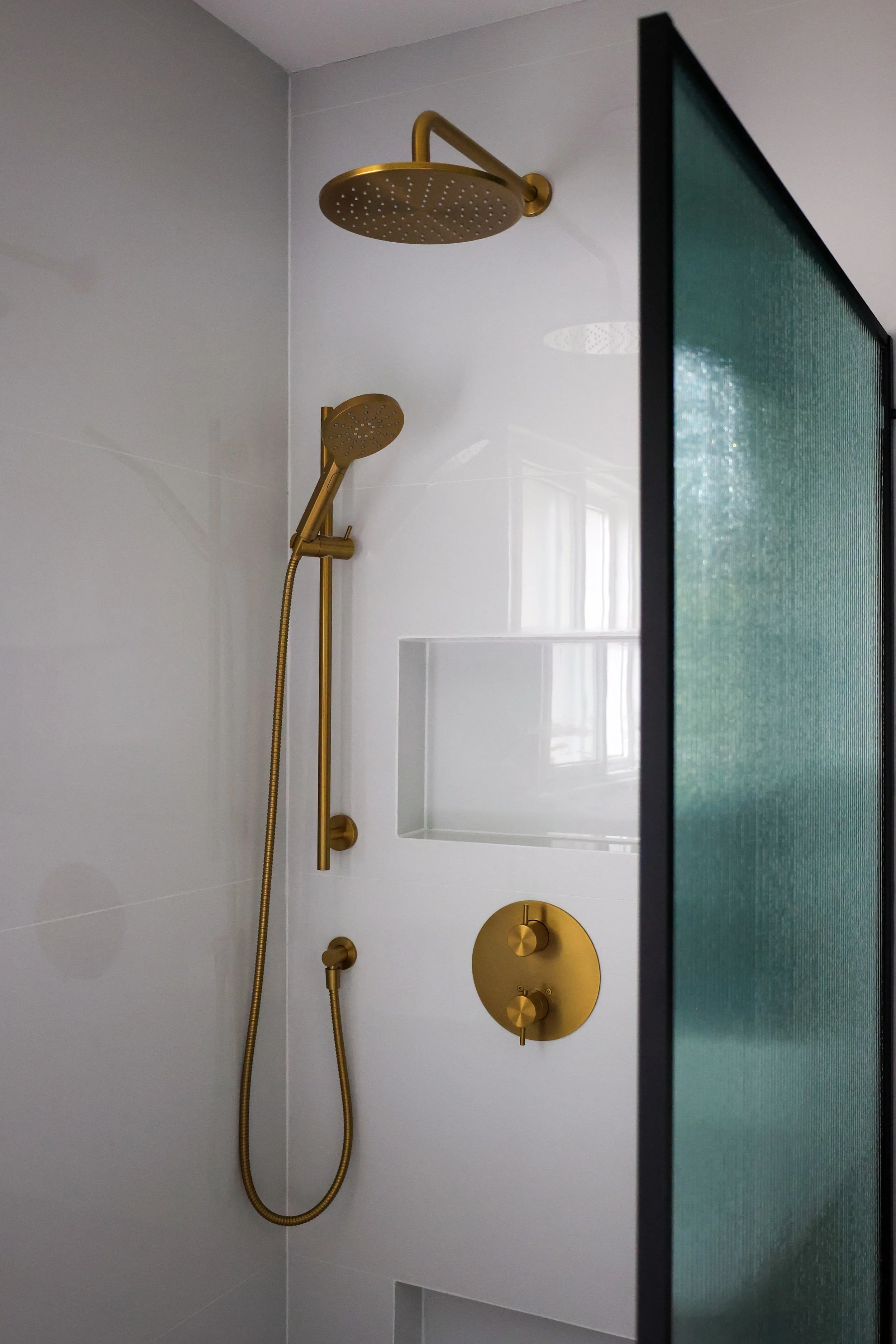 Modern shower with wall-mounted gold fixtures, including a large overhead showerhead and a handheld showerhead, behind a black-framed glass screen.