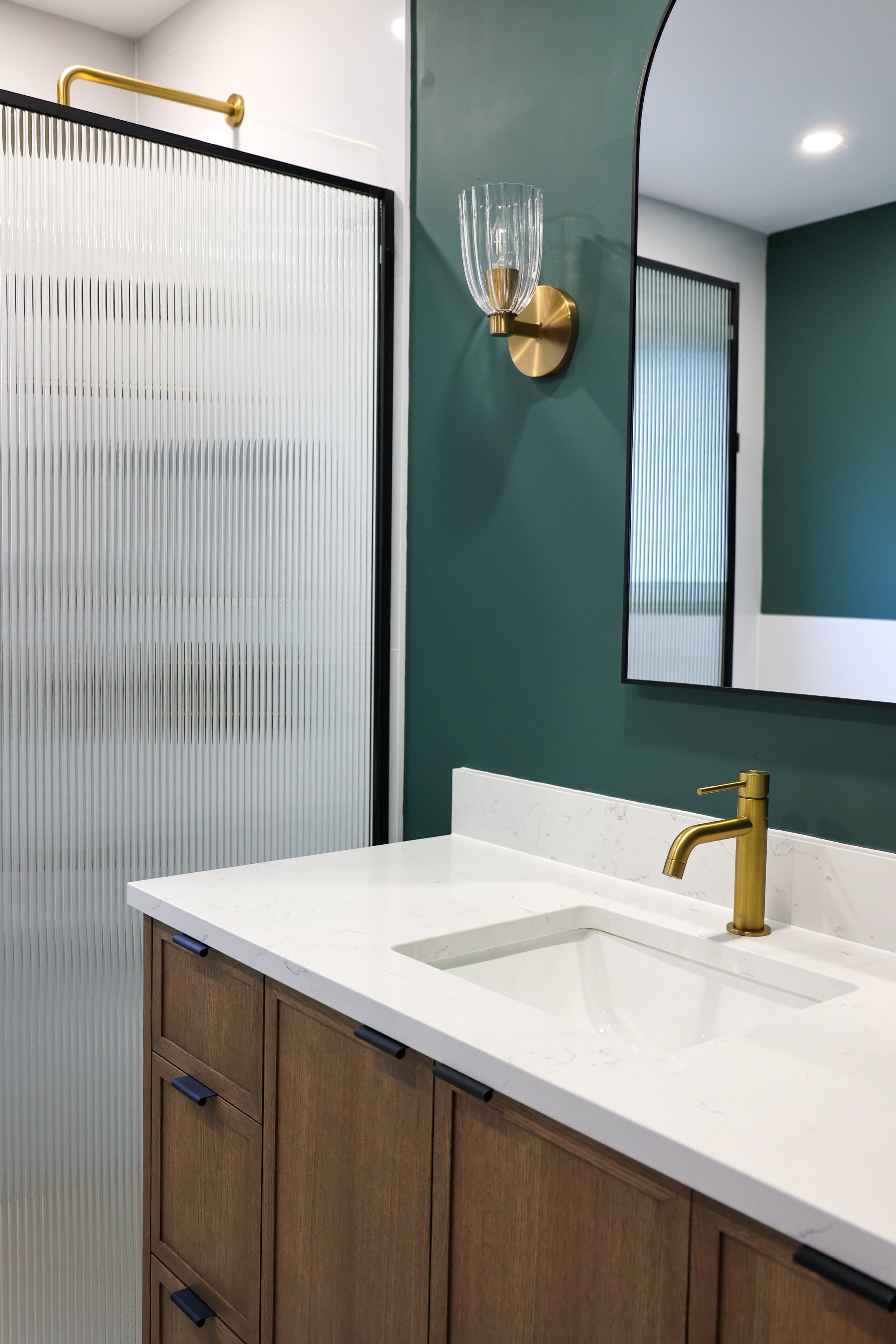 Bathroom vanity with white marble countertop, wooden cabinets, gold faucet, mirror, and green wall with a modern wall sconce, next to a frosted glass shower door.