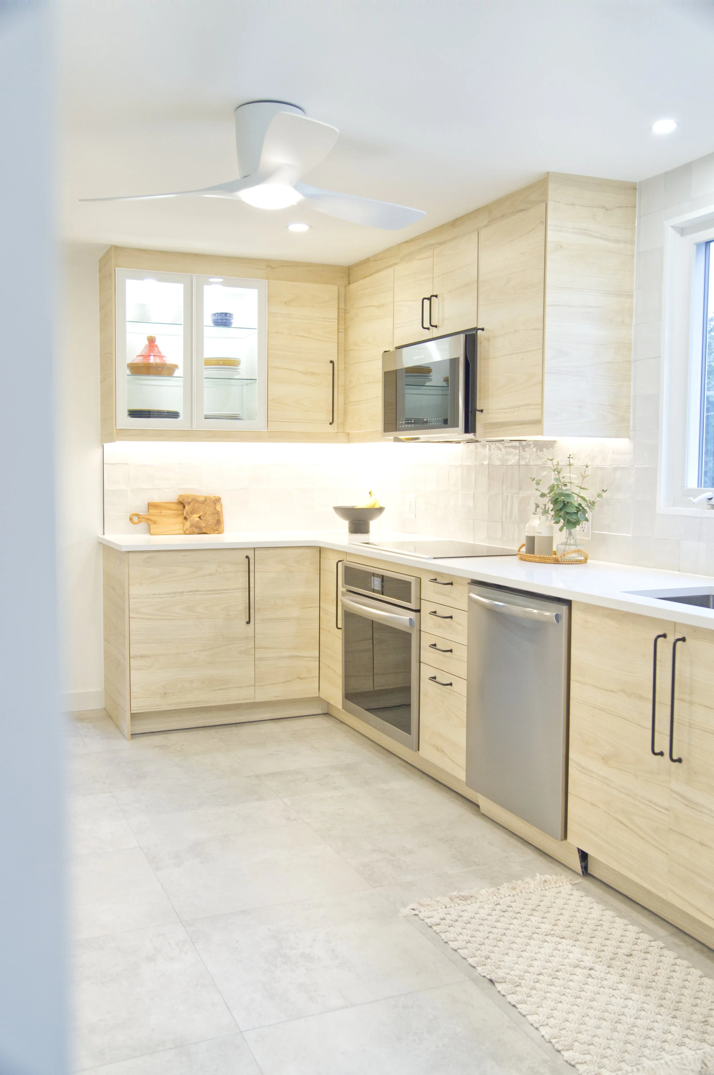 Modern kitchen with light wood cabinets, white countertops, stainless steel appliances, and a window with natural light.