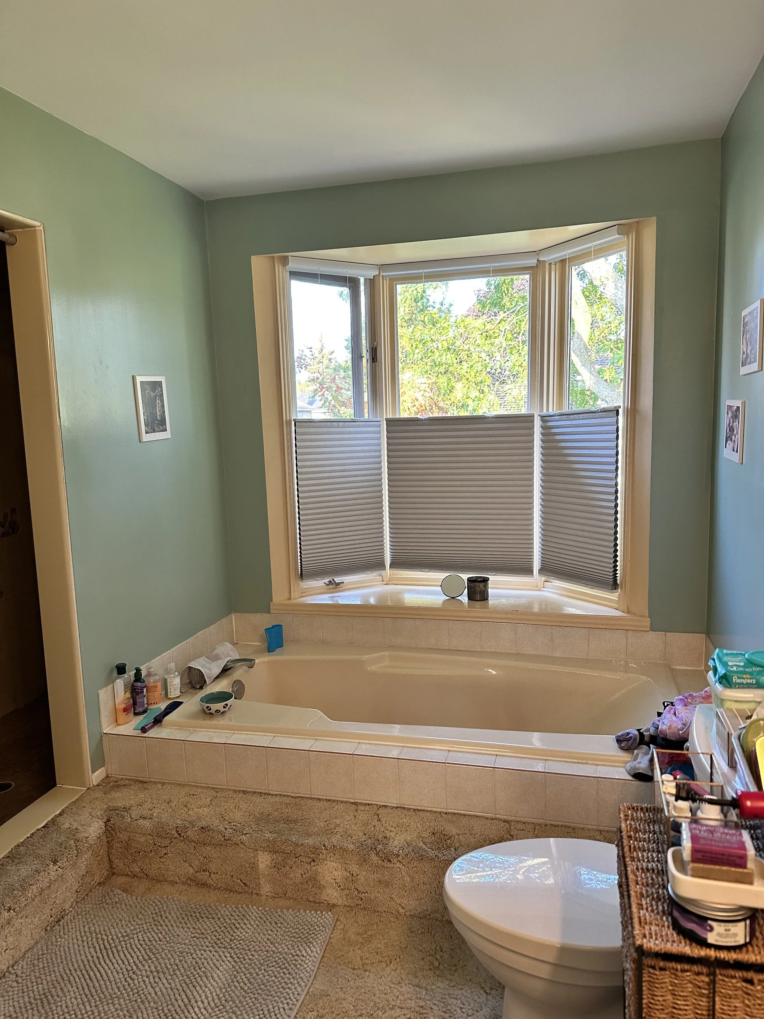 A bathroom featuring a bathtub by a bay window with partially closed blinds, a toilet, and a shelf with toiletries.