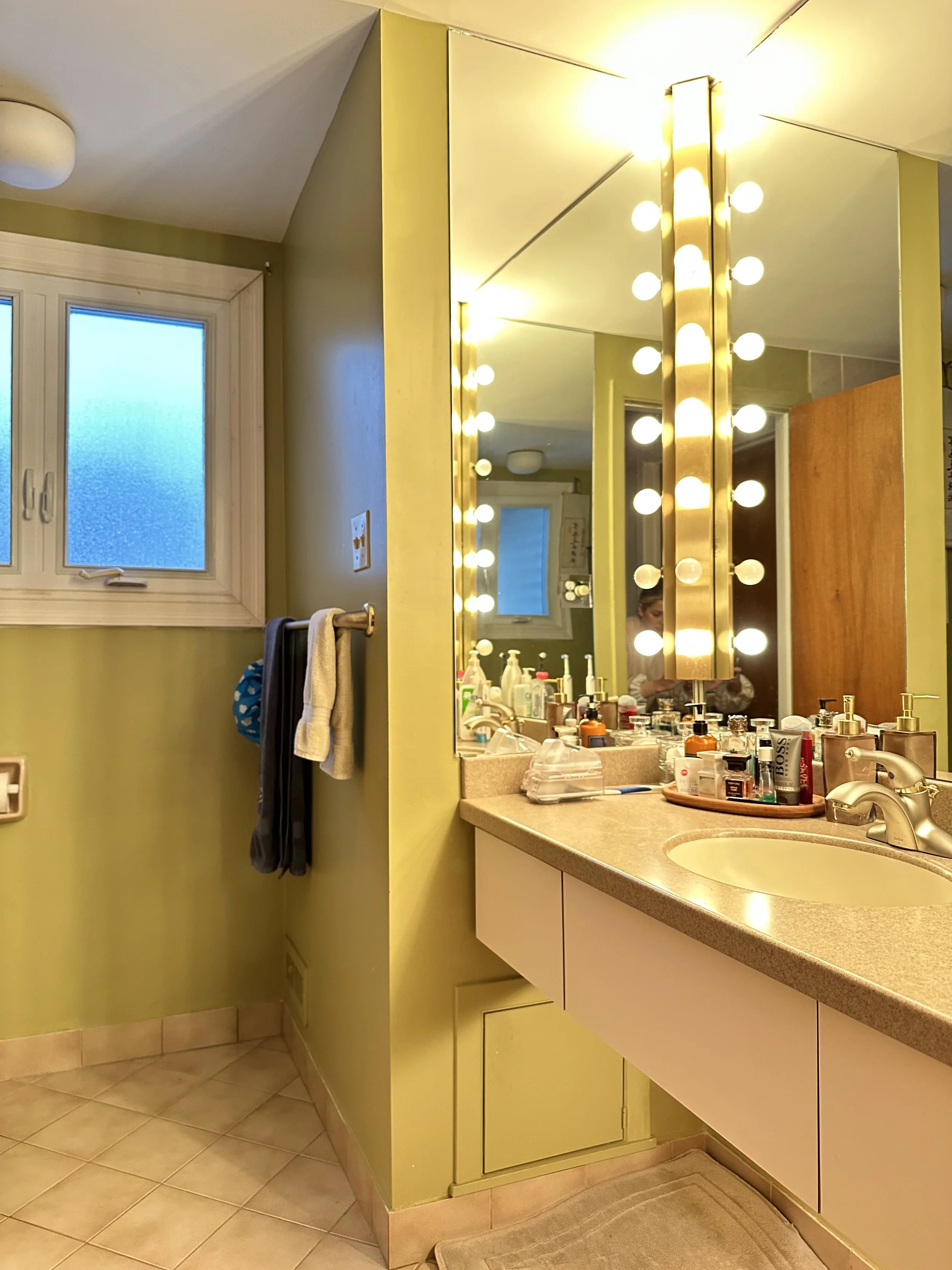 Bathroom with a green wall, a window, a mounted towel rack with towels, a large mirror with vanity lights, a beige countertop with various toiletries, and a sink.