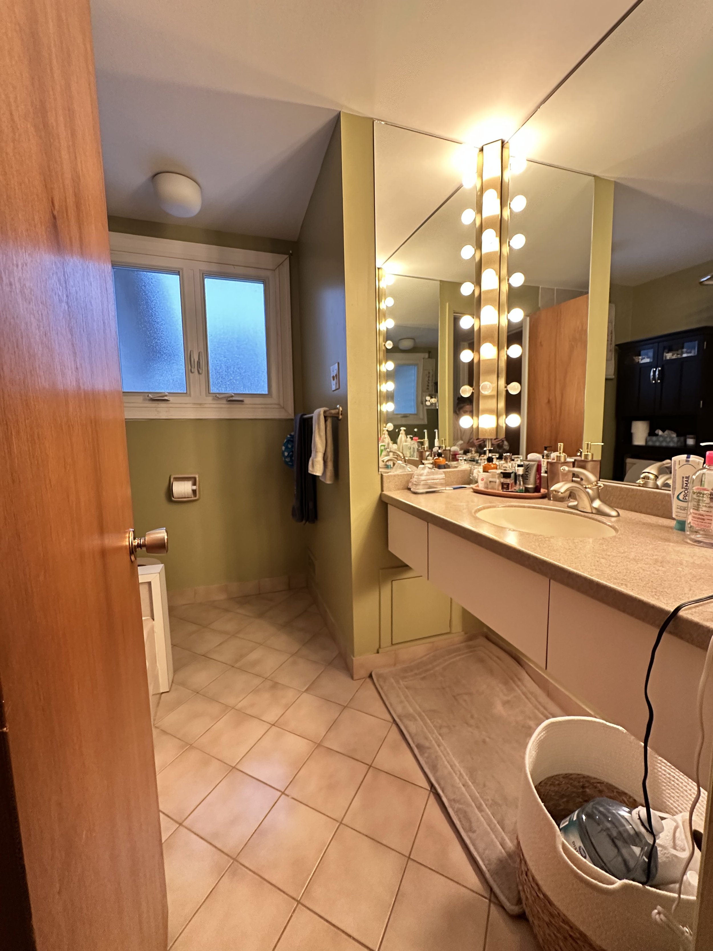 Bathroom with large mirror framed by lights, beige countertop with toiletries, green walls, tiled floor, window, and laundry basket.