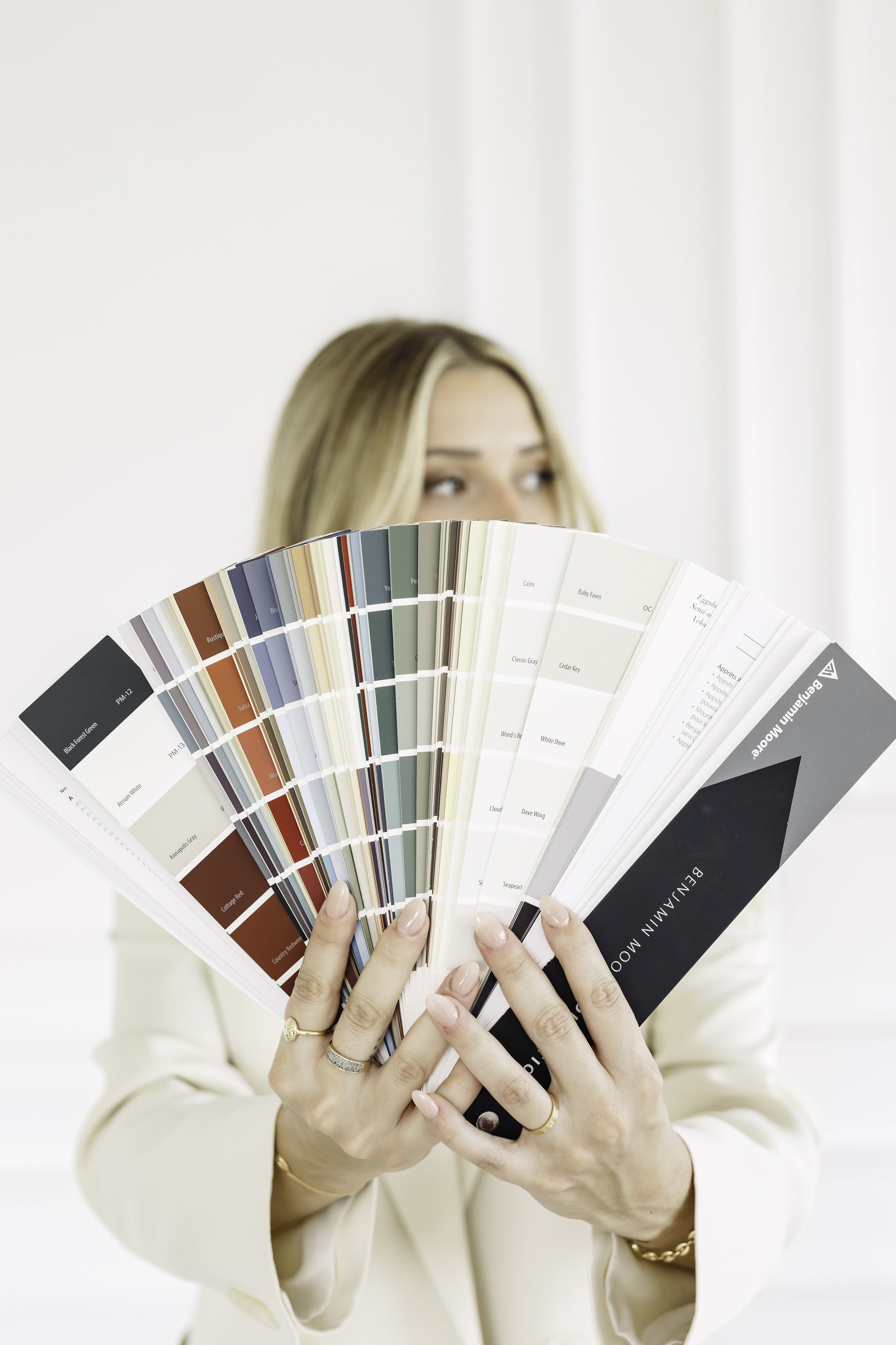 Woman holding a color swatch book in front of her face, showing various paint colors.