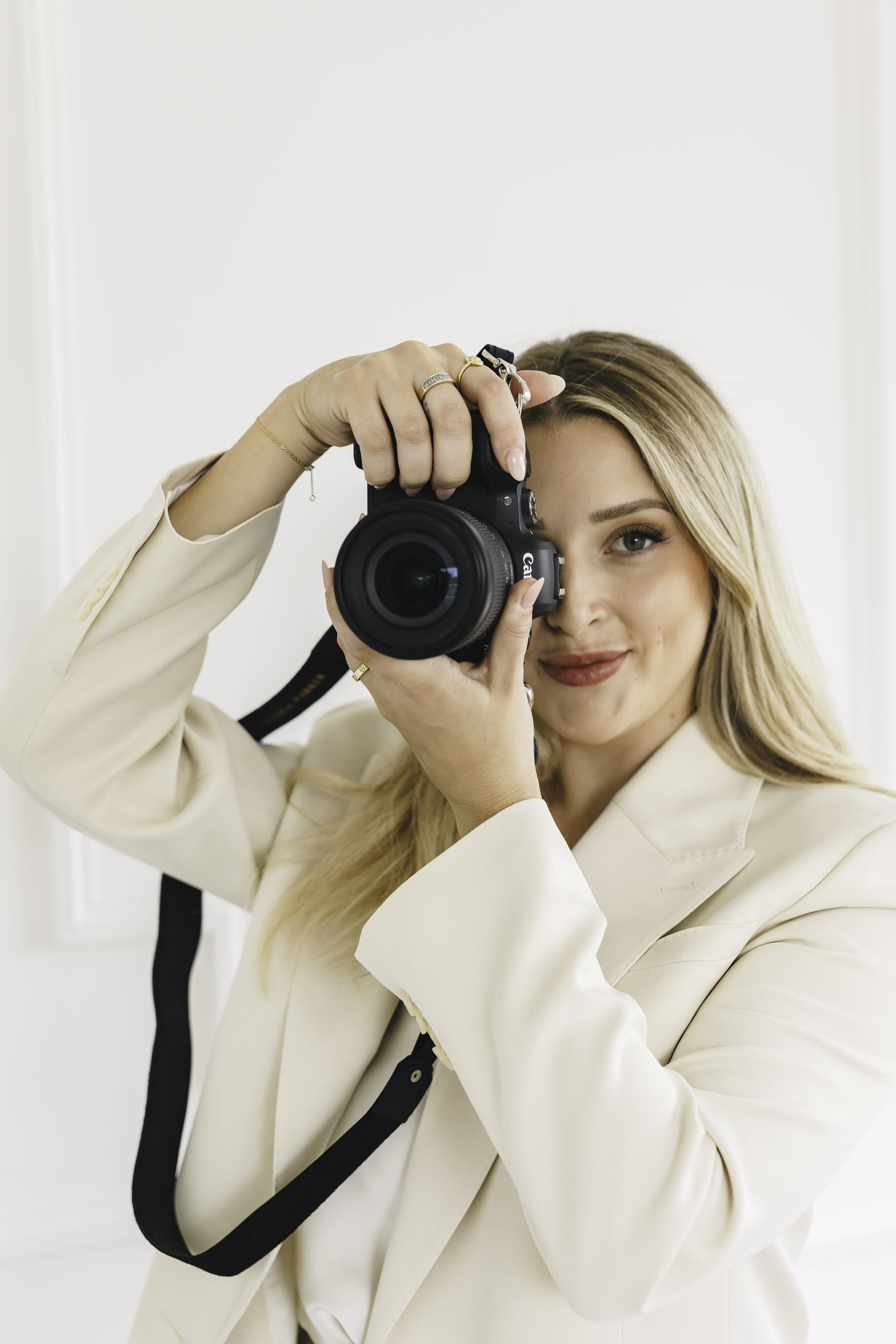 A woman with blonde hair and a beige blazer is holding a camera, taking a photo of herself in a mirror.