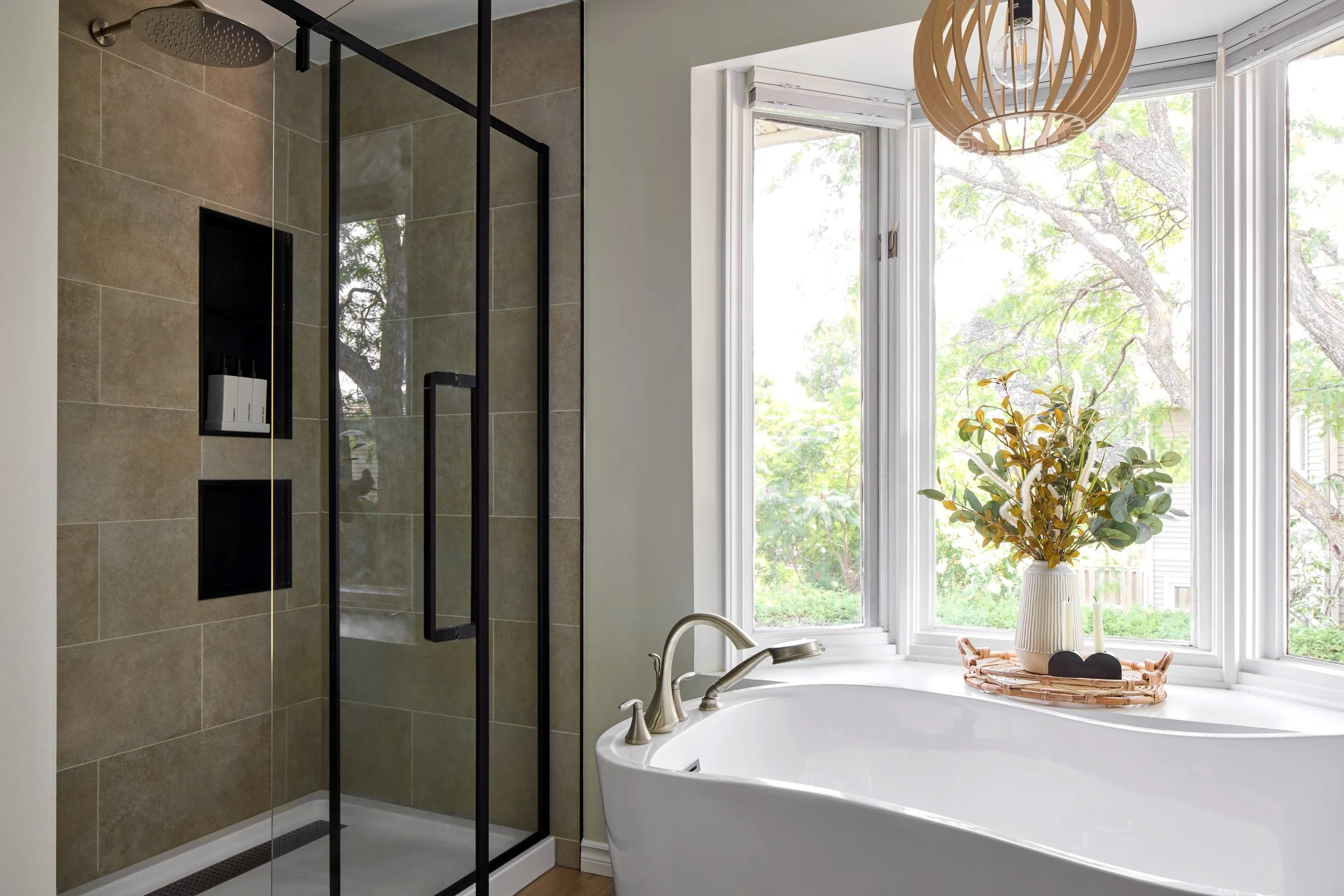 Bathroom with a walk-in shower with glass door and black frame, beige tile walls, a white bathtub next to large bay window with greenery outside, and a hanging decorative wooden light fixture.