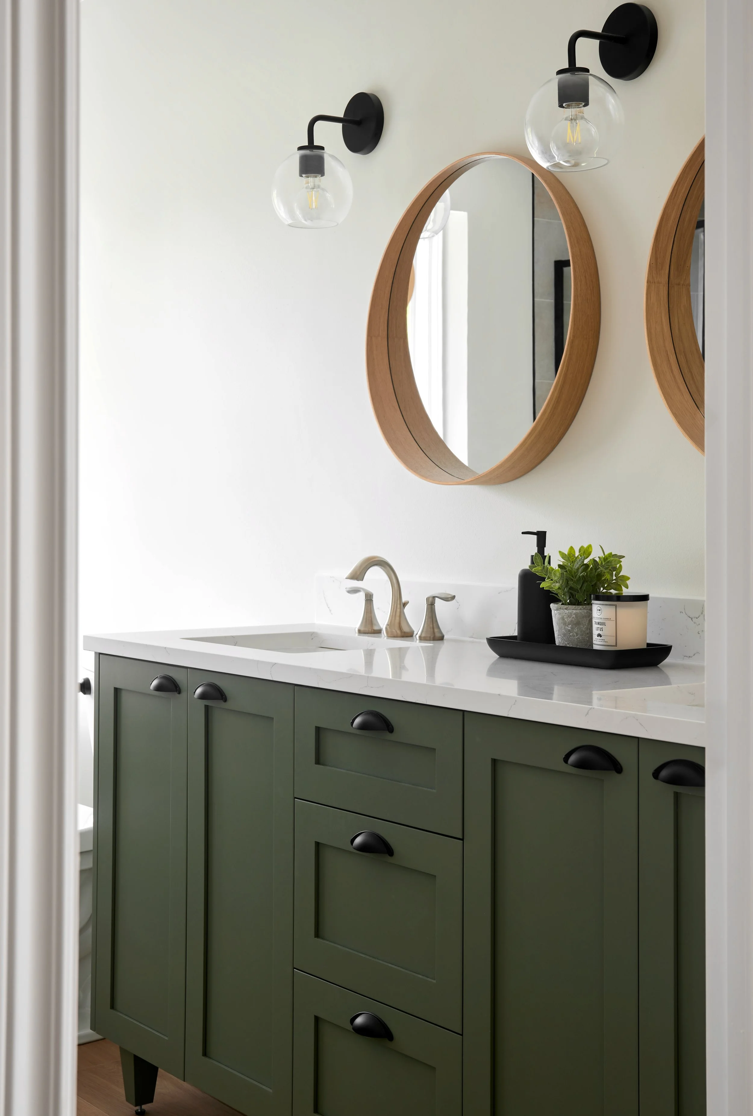 Bathroom with green vanity, white marble countertop, oval mirror with wooden frame, black light fixtures, and decorative items including a black soap dispenser and a potted plant.