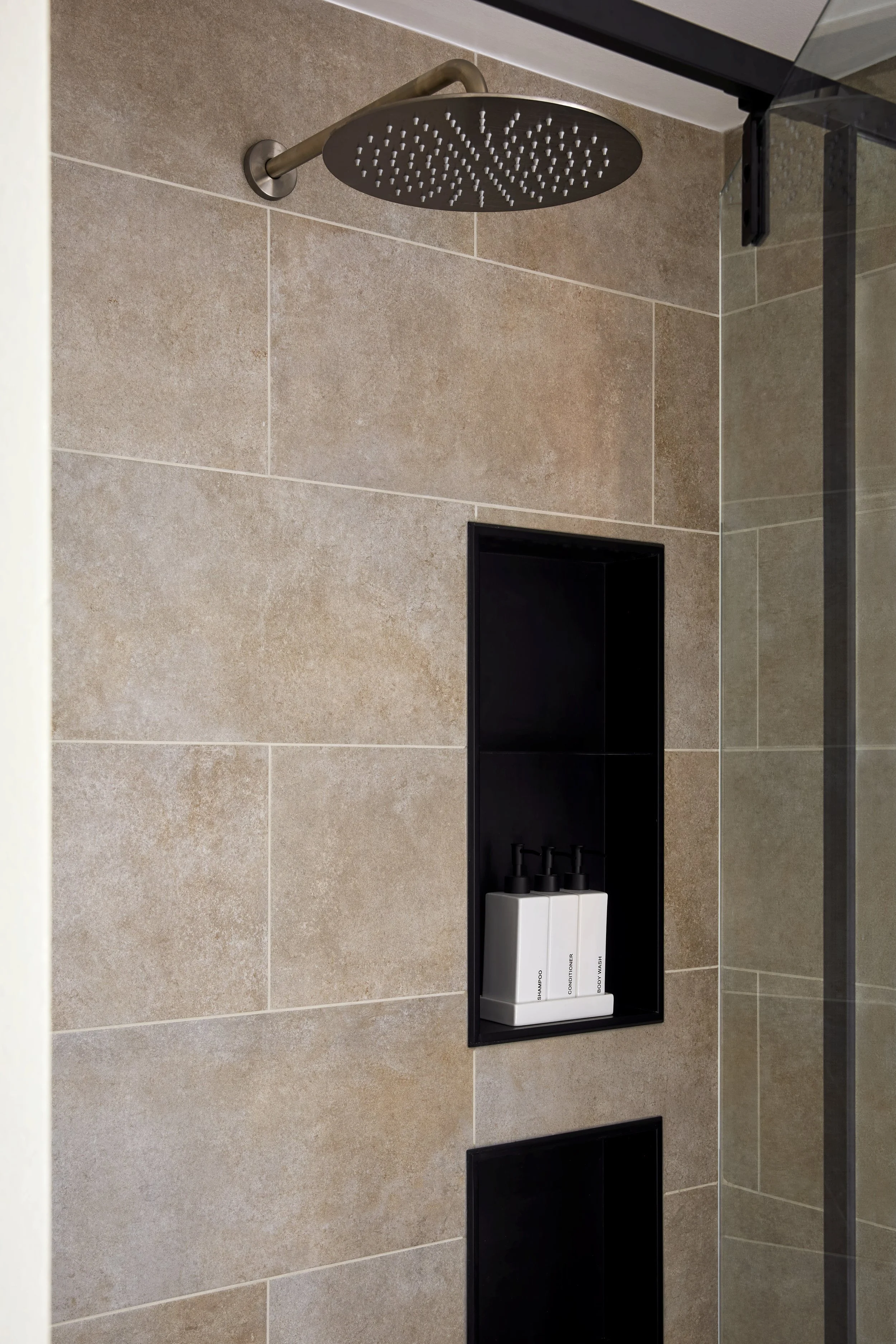 Close-up of a shower wall with beige tiles, a black recessed shelf holding three white bottles with black pumps, a black vent below, and a partial view of a glass shower door.
