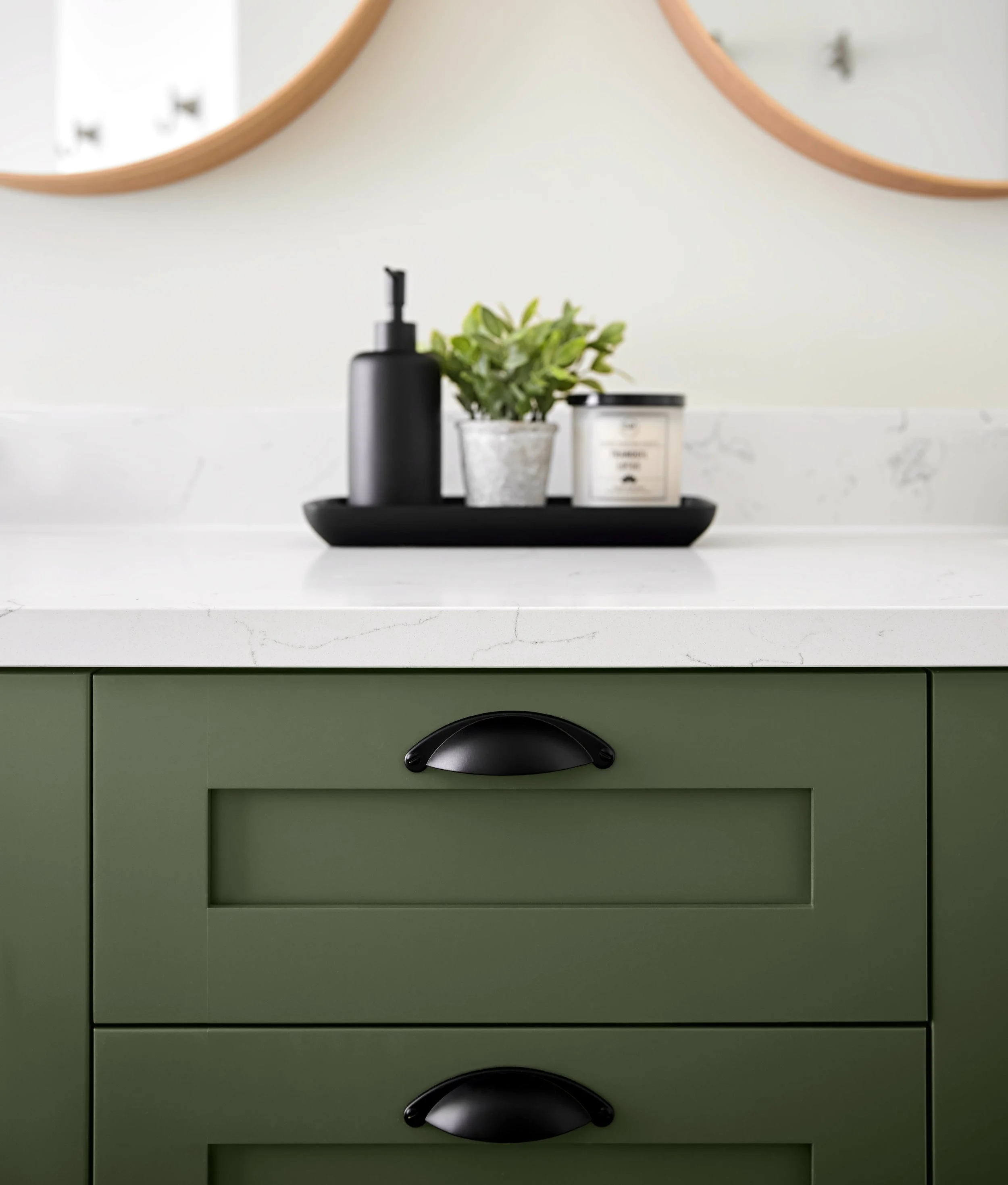 Green kitchen cabinet with black handles, white marble countertop, black soap pump, potted green plant, black tray holding a white candle in a jar, two octagonal mirrors with wooden frames on the wall behind.