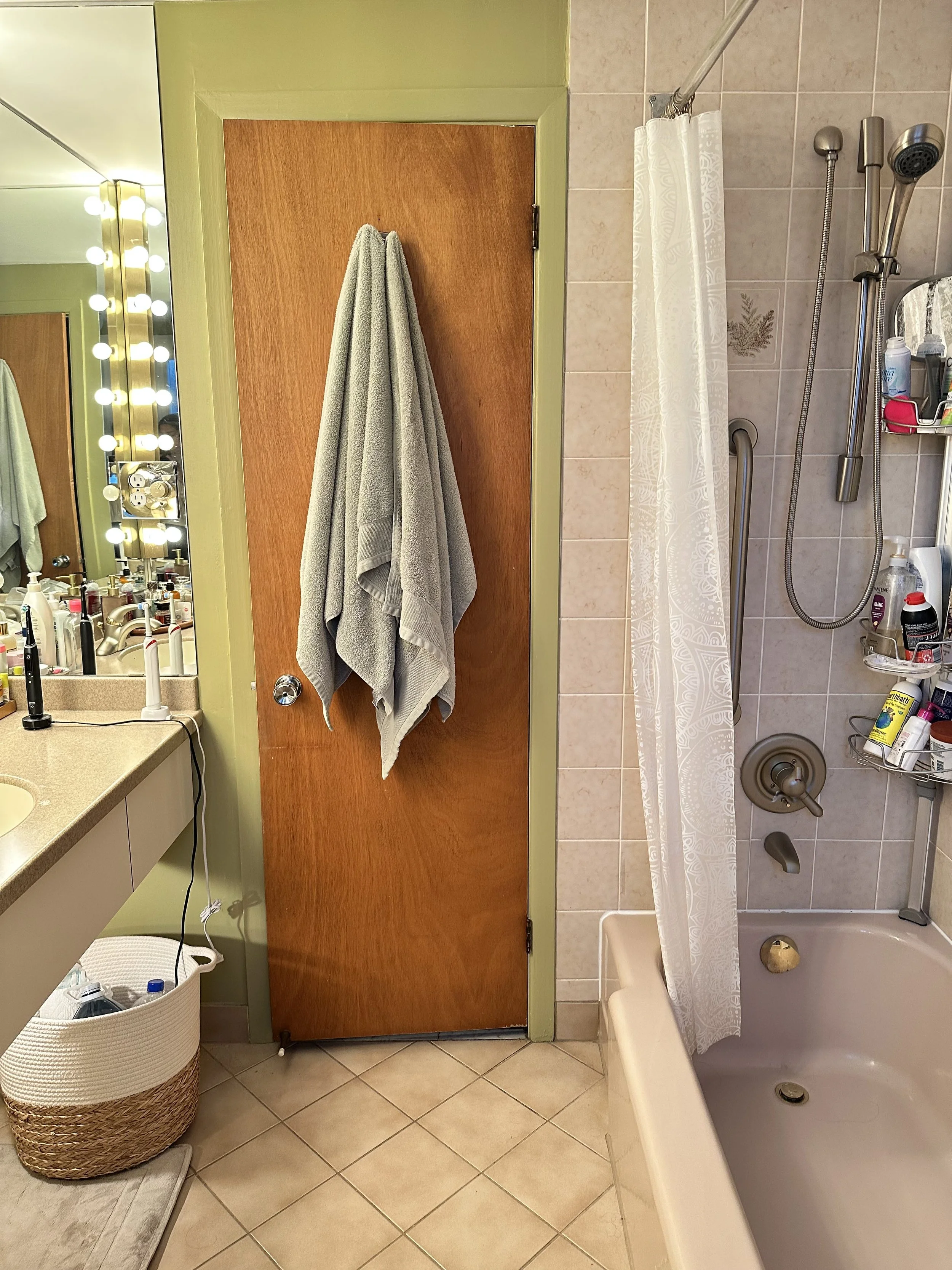A bathroom with a wooden door, a towel hanging on it, a mirror with light bulbs, a countertop with toiletries, a laundry basket, and a bathtub with a shower curtain and shelves.