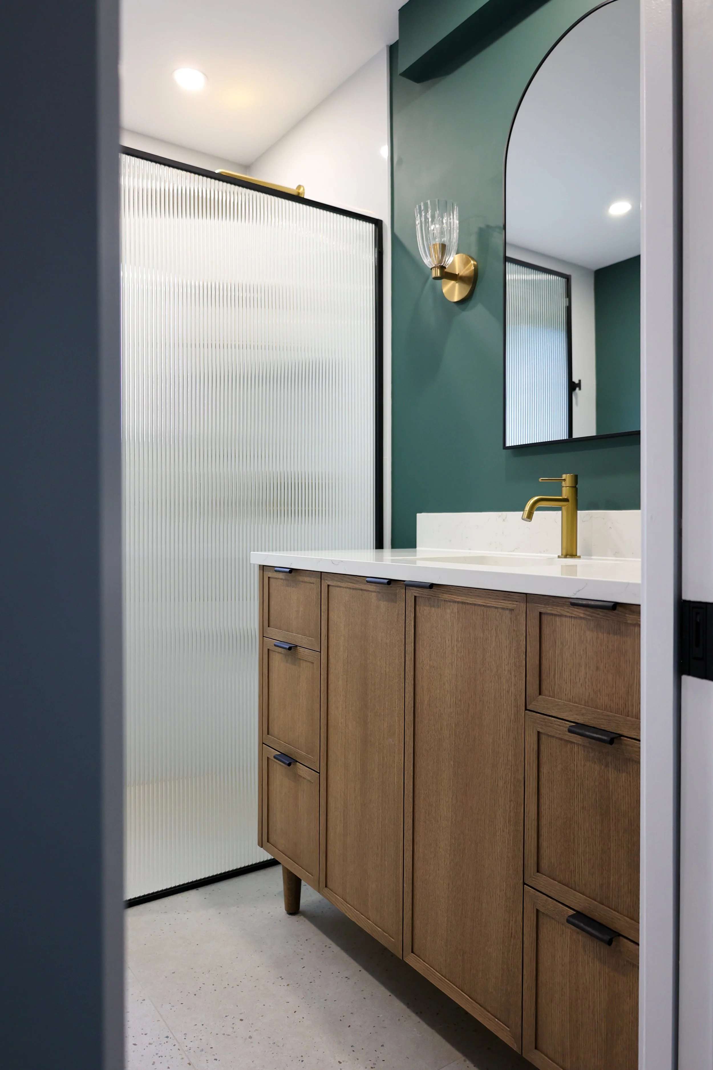 Modern bathroom with green accent wall, wooden vanity with black handles, white countertop, gold faucet, and a large mirror. A gold wall sconce with a glass shade is on the wall, and a frosted glass shower door is visible.