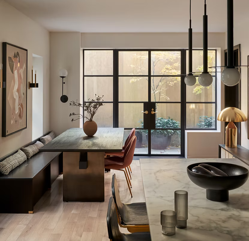 Modern dining room with a large table, surrounded by chairs, featuring a pendant light fixture, large windows with a view of a patio, and contemporary decor including artwork, a vase with branches, and a table lamp.